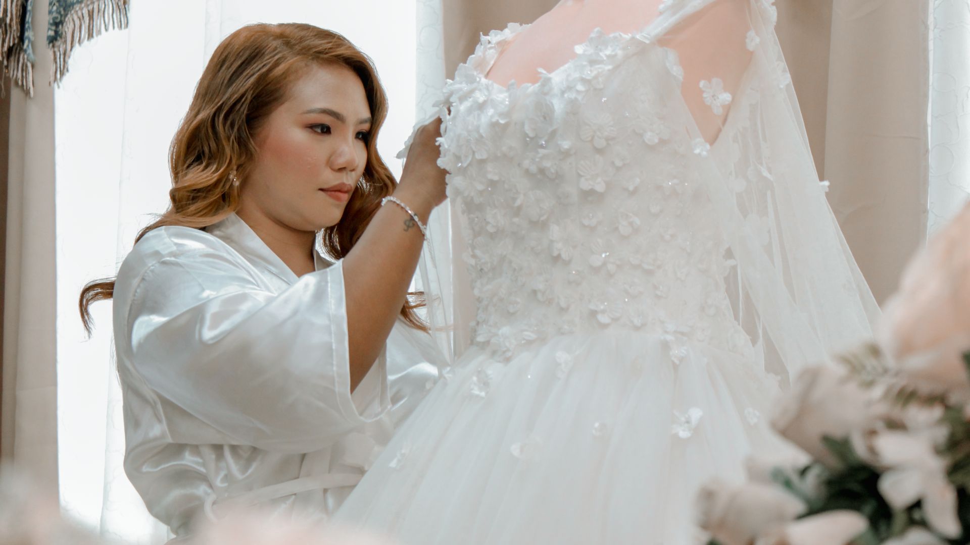 Woman in white robe carefully adjusts detailed wedding dress indoors.