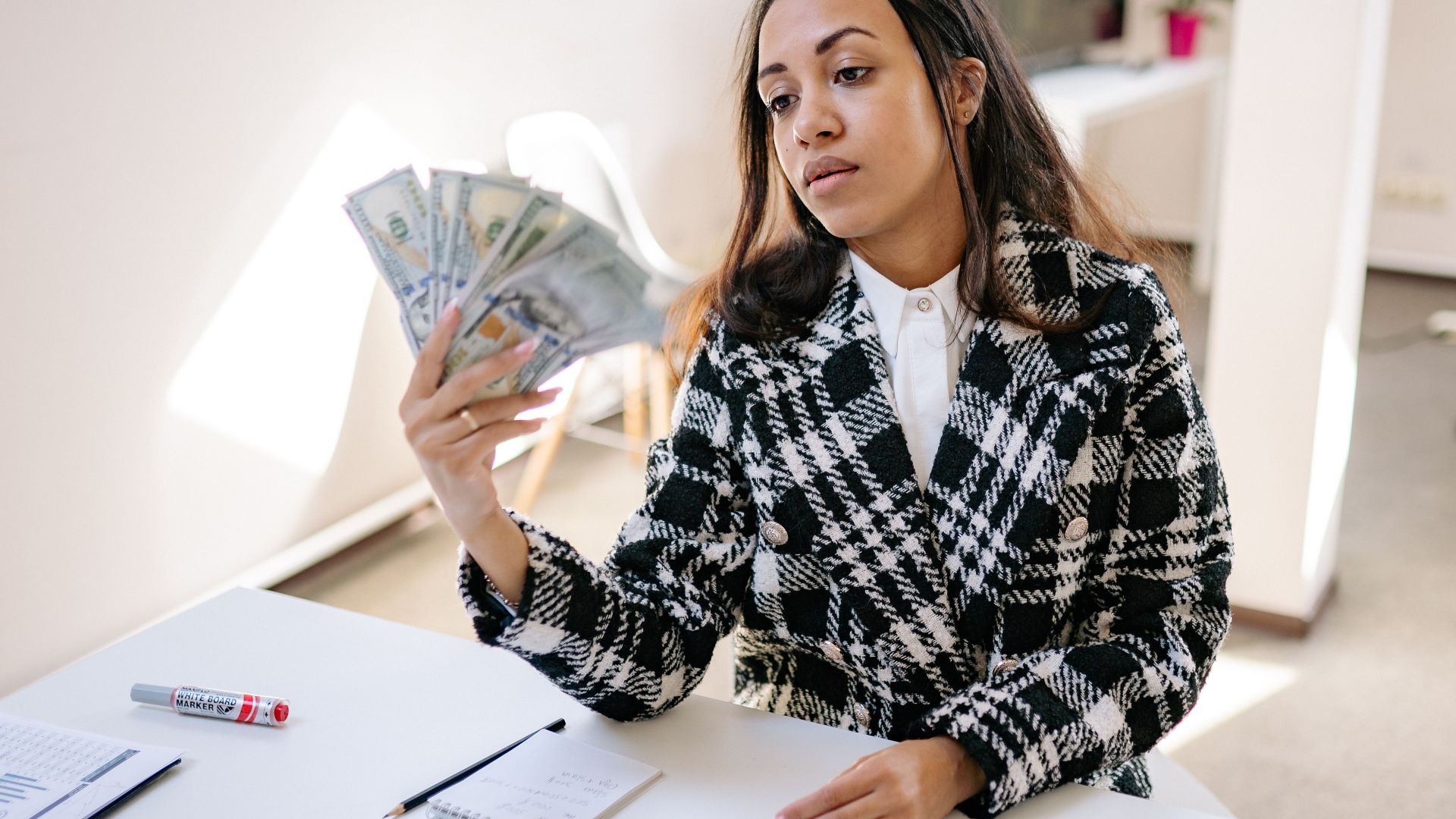 A young woman in a plaid coat counting US dollar bills at a desk in a modern office.