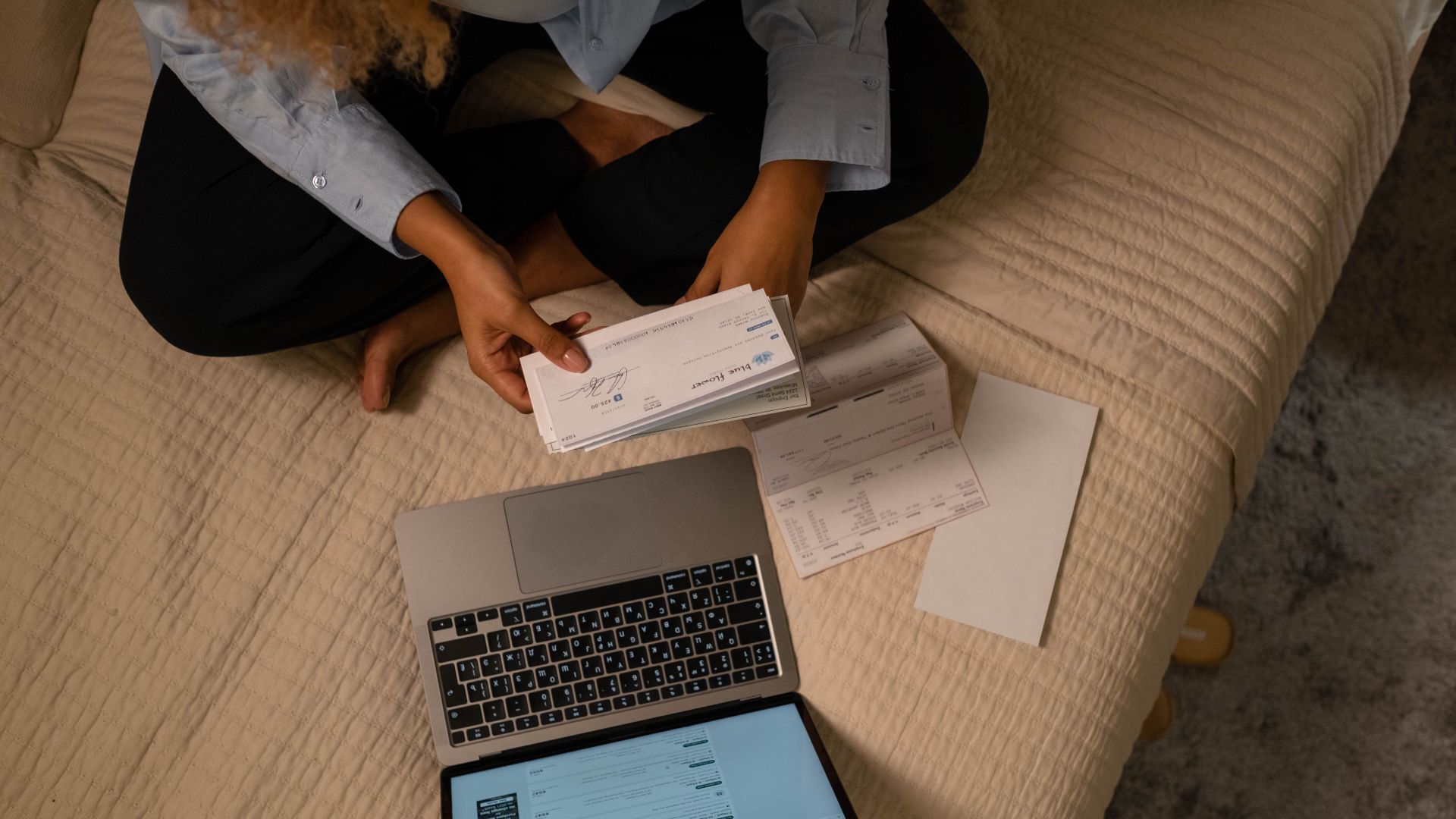 Overhead view of woman organizing finances on bed with laptop and checks.