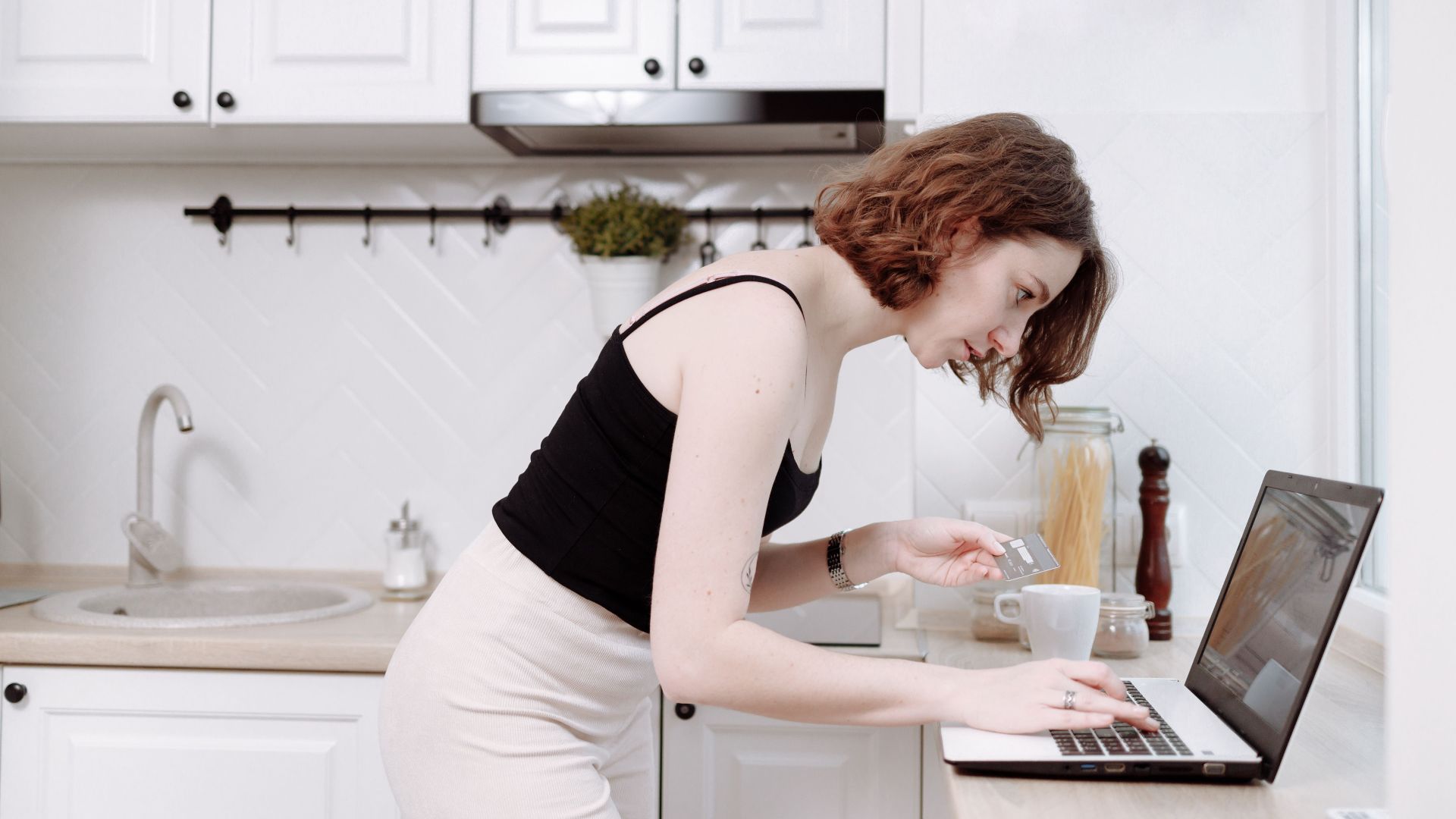 Young woman using a laptop in a kitchen, making an online purchase with a credit card.
