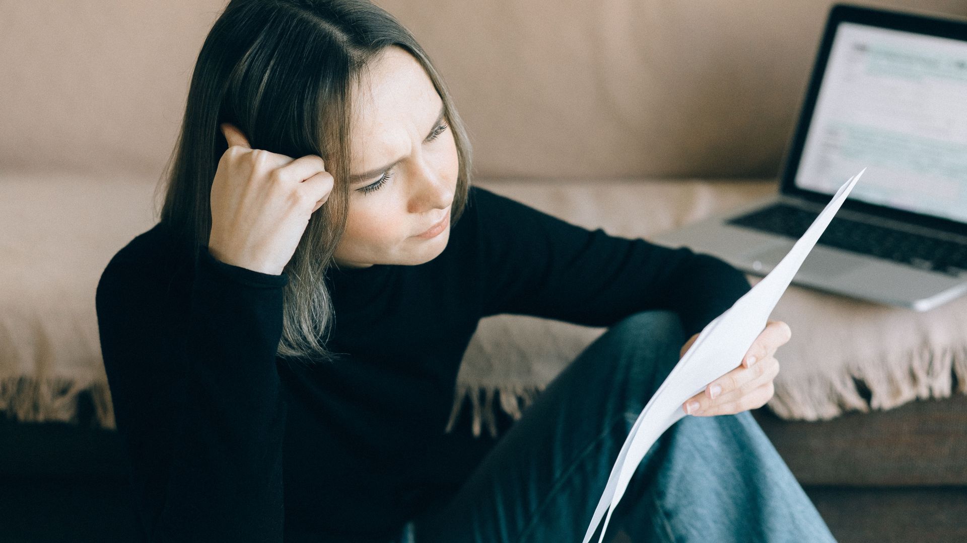 Focused woman analyzing papers with a laptop open, symbolizing thoughtful consideration.