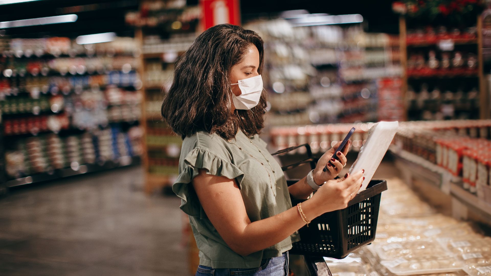 A woman shopping in a supermarket, wearing a face mask and using her phone to check products.