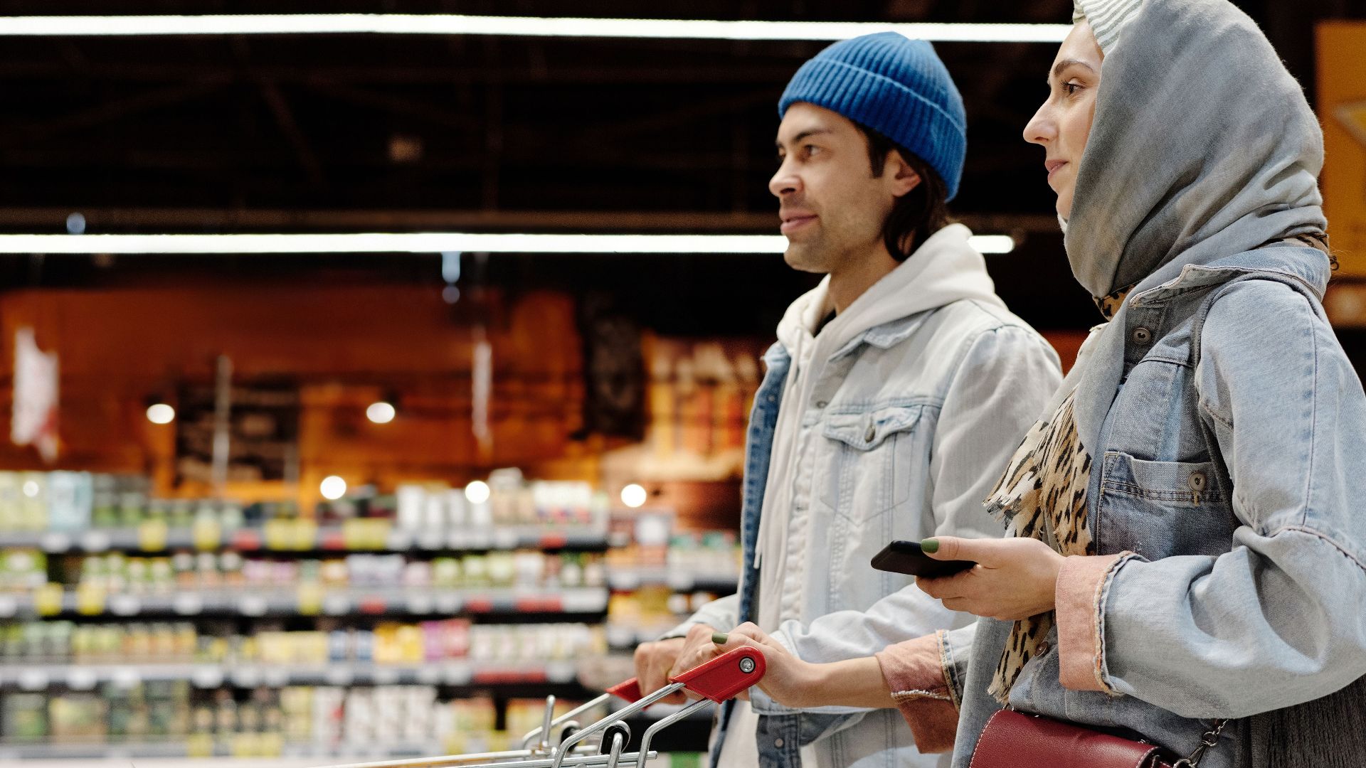 A young couple shopping with a cart in a supermarket aisle, selecting fresh produce.