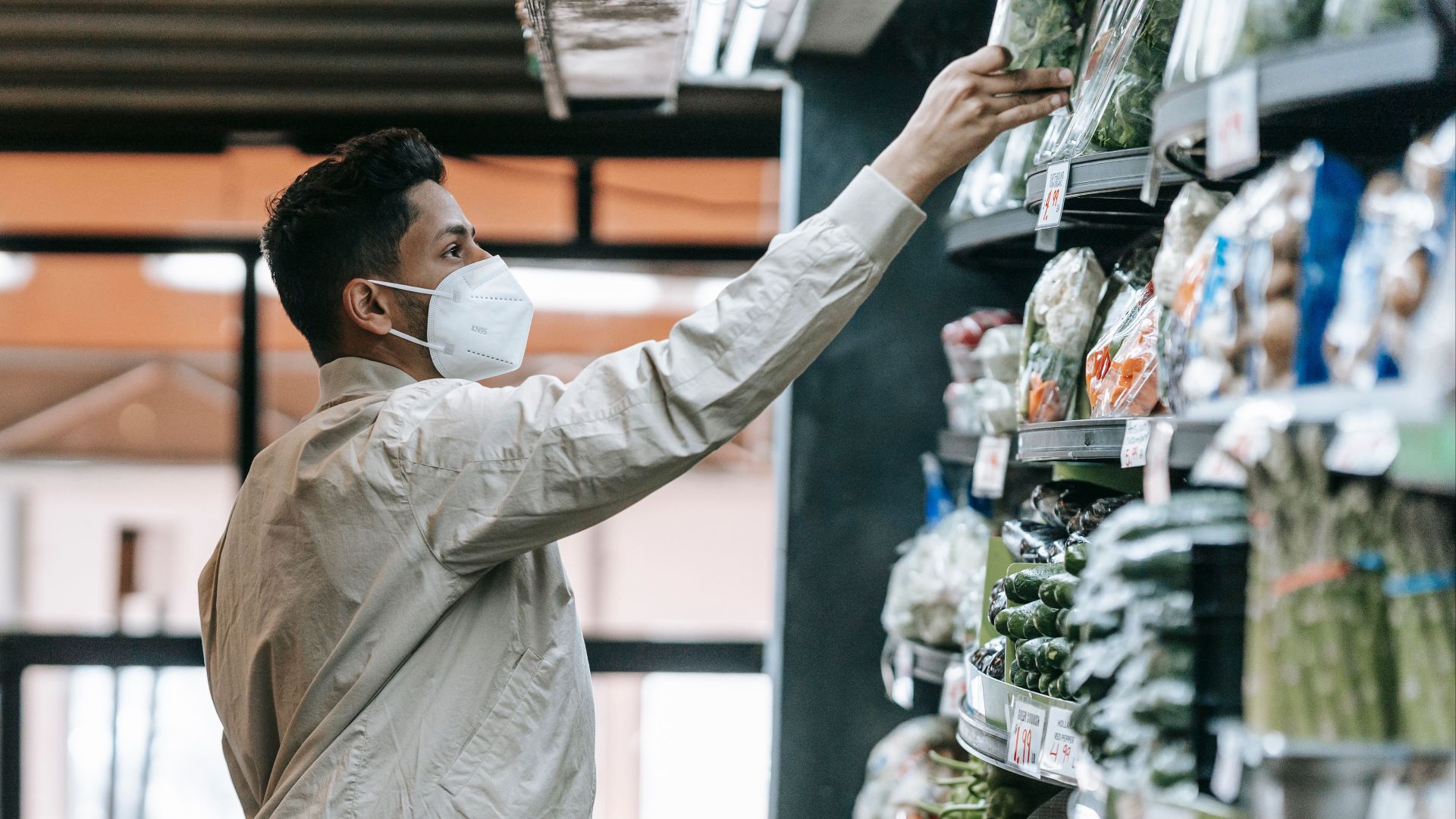 Side view of Indian male customer in protective mask picking fresh products from stall while shopping in supermarket during pandemic