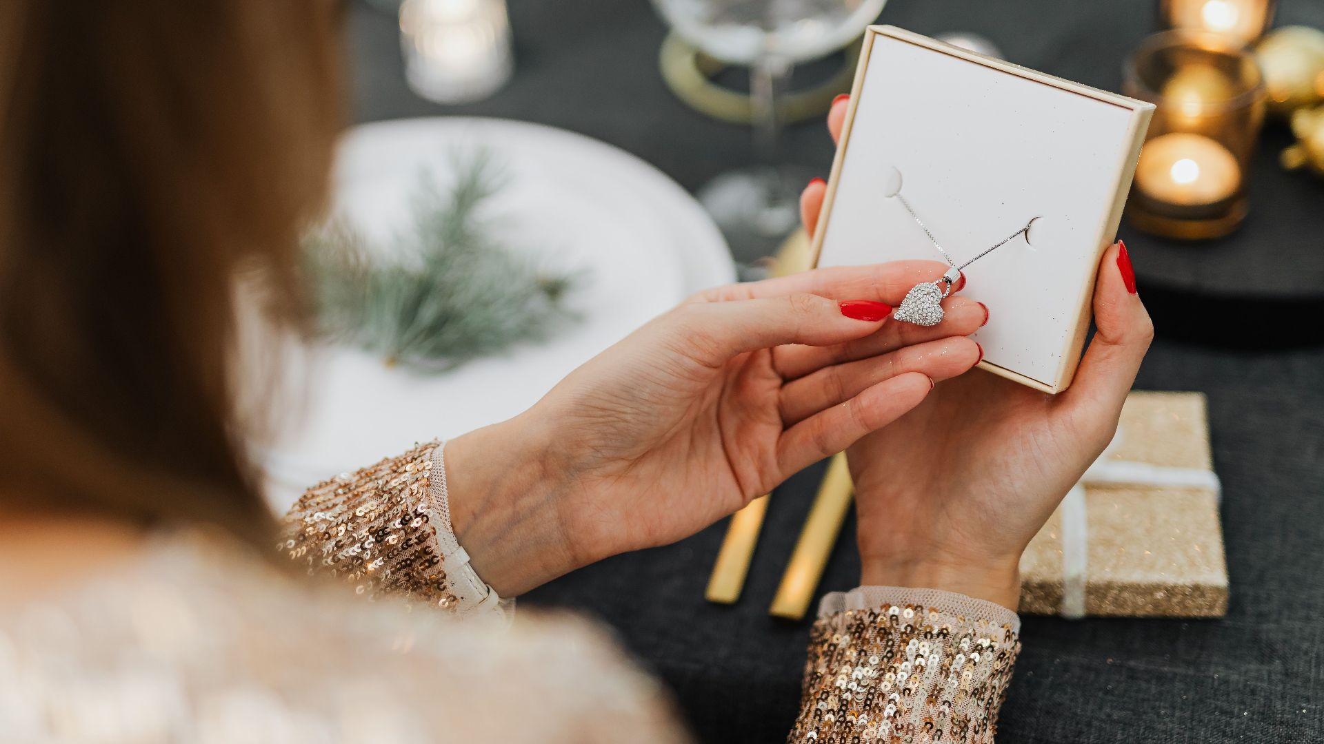 A woman opens a necklace gift surrounded by a festive dinner setting, creating a warm, celebratory mood.