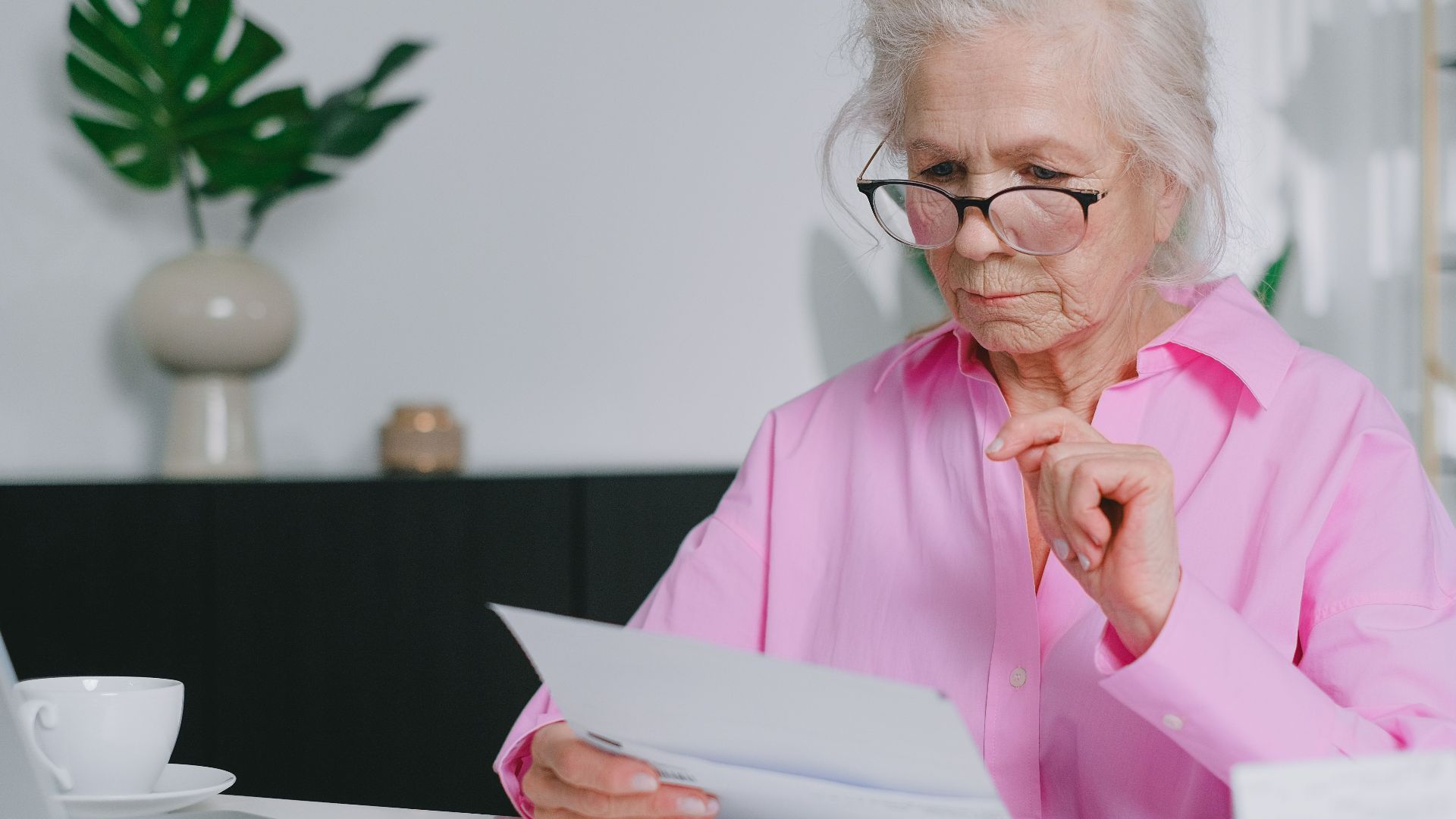 Elderly woman in pink shirt reading documents at home workspace.