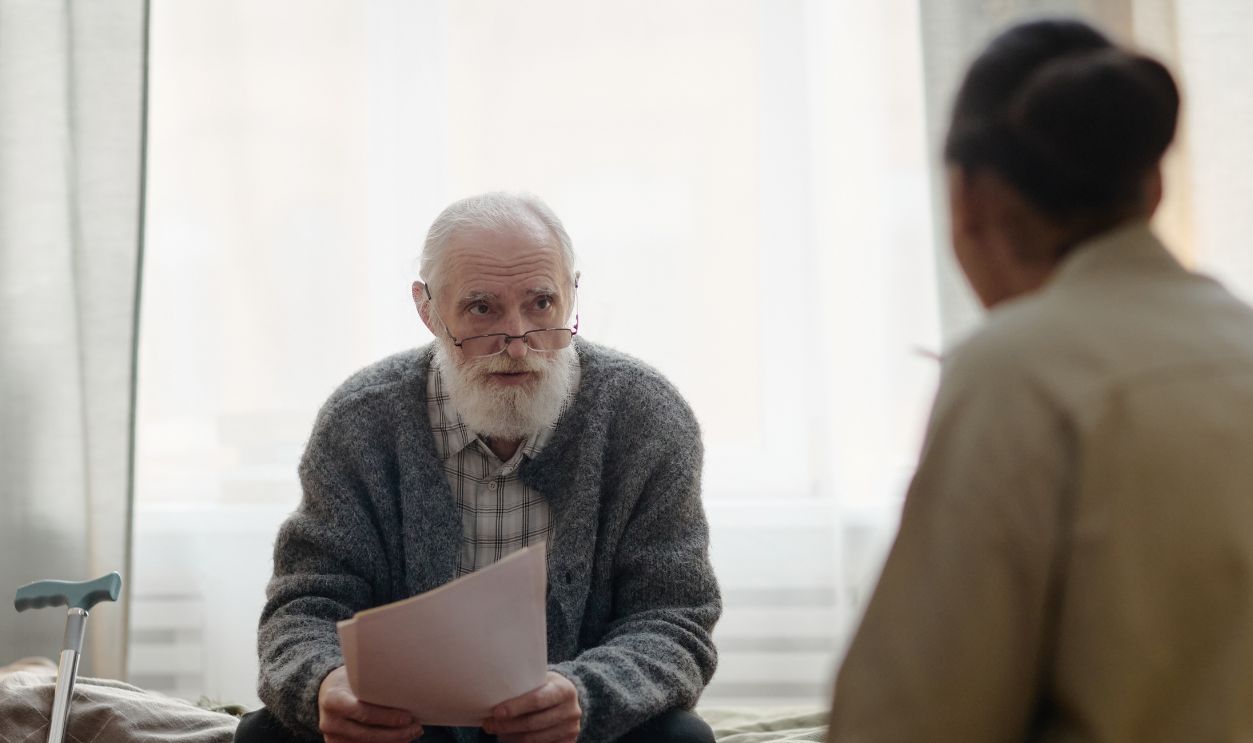Senior Caucasian man sitting on bed holding documents talking with middle aged Black woman social worker in home setting, walking cane visible beside bed, both engaged in conversation