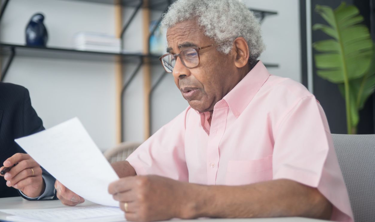 Elderly man with glasses reading a document in a home office setting, reflecting focus and concentration
