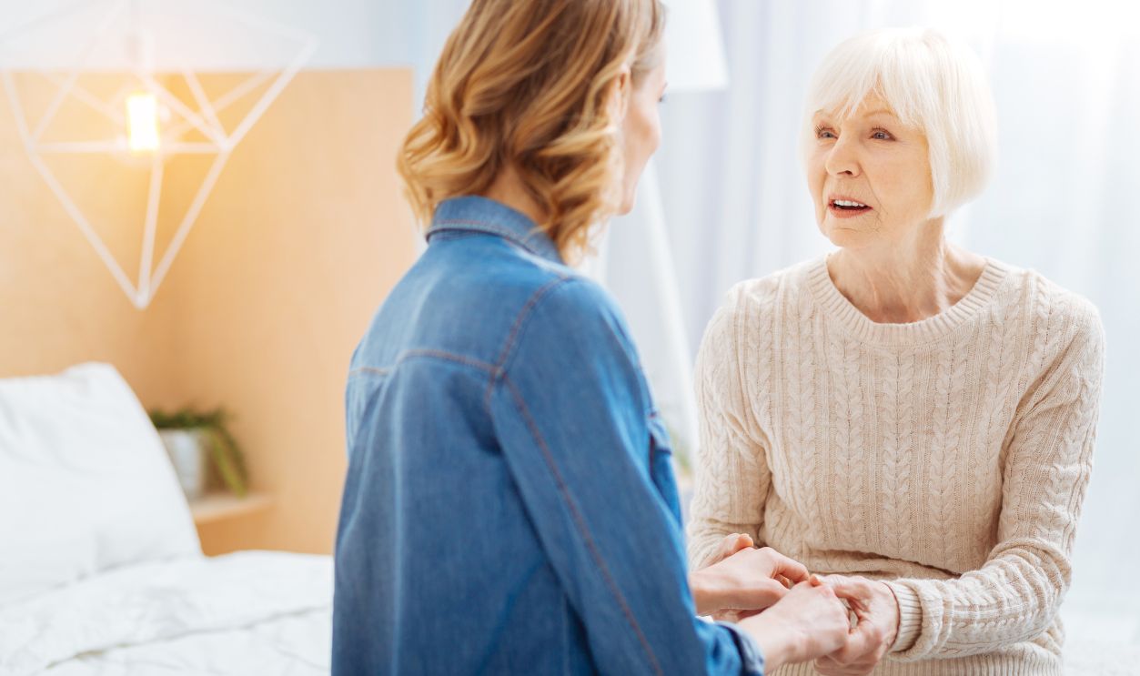Shutterstock-1043238988, Urgent talk. Emotional senior lady sitting on a soft bed in front of her kind young granddaughter and looking at her while touching her hands and talking about an urgent problem