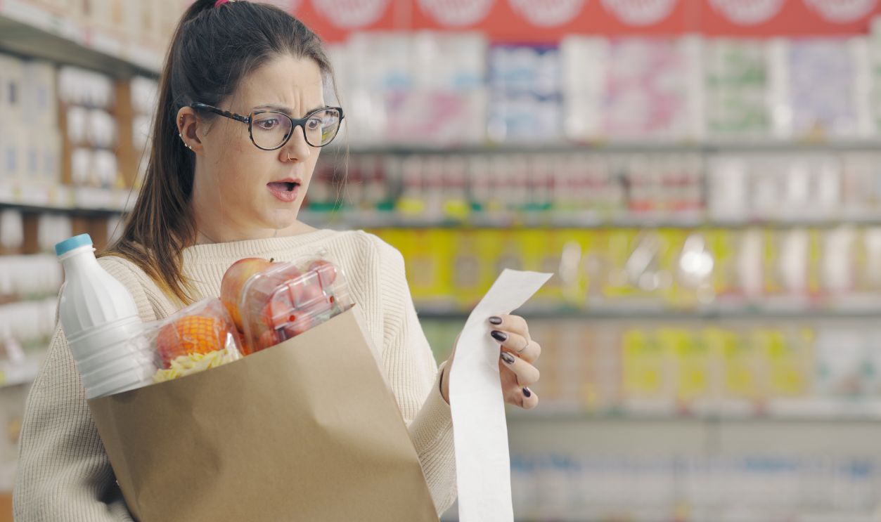 Shutterstock-2387422431, Shocked woman checking a long grocery receipt at the supermarket, expensive grocery and inflation concept