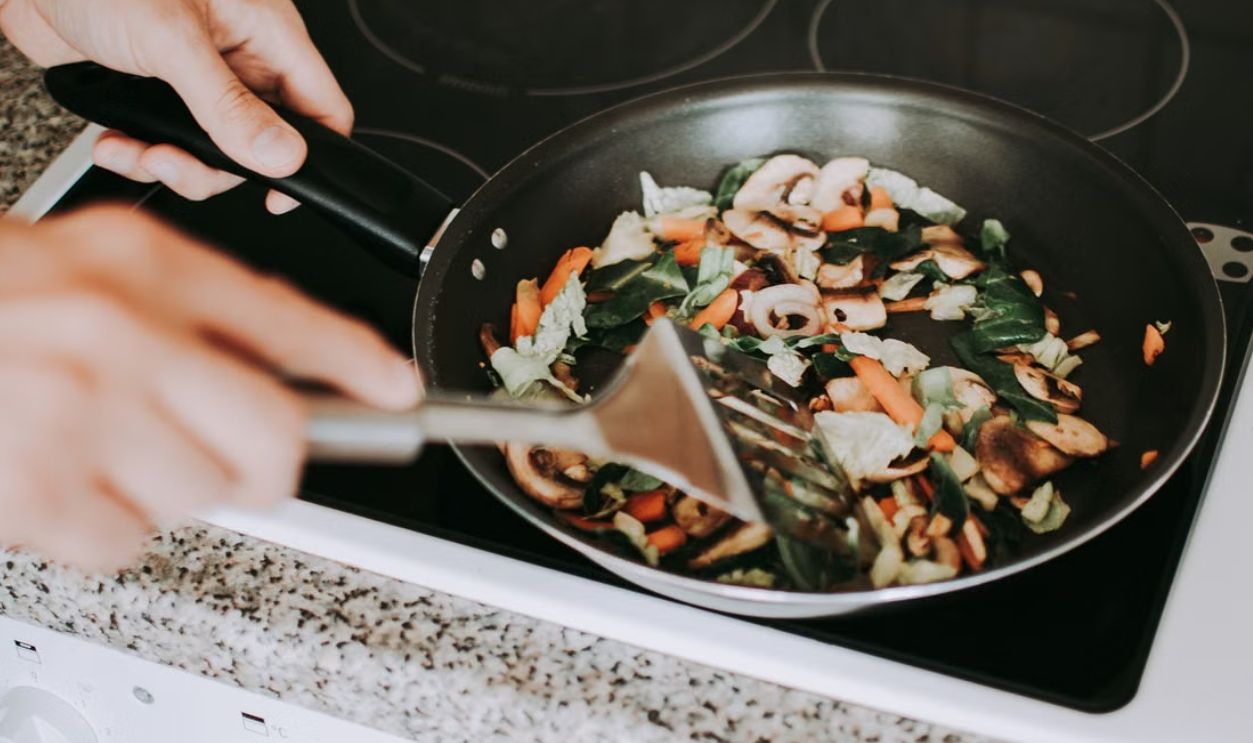 A person stirring vibrant vegetables in a frying pan on an electric stove