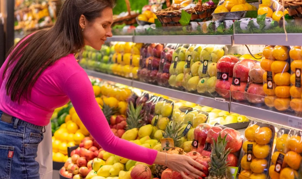 A woman in a bright pink top selecting fresh fruits in a grocery store produce aisle