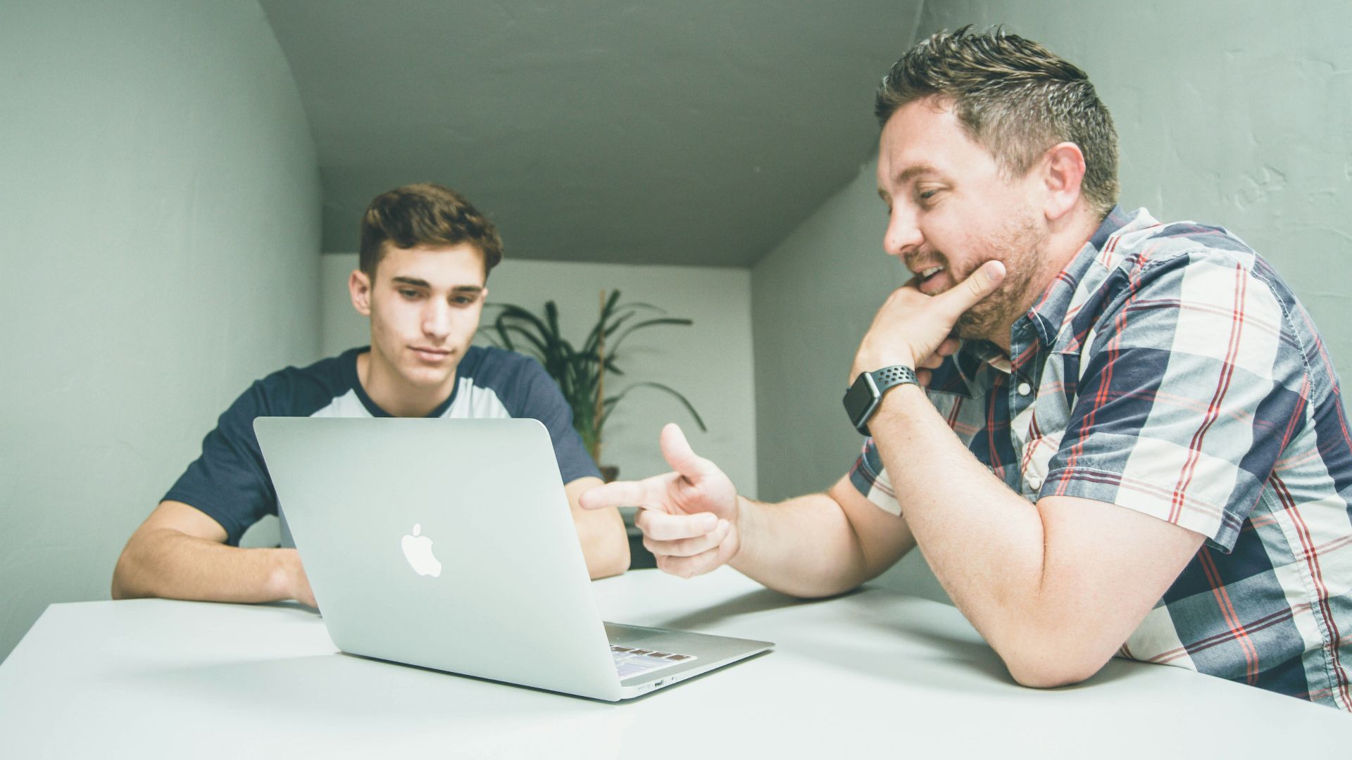 man wearing white and black plaid button-up sports shirt pointing the silver MacBook