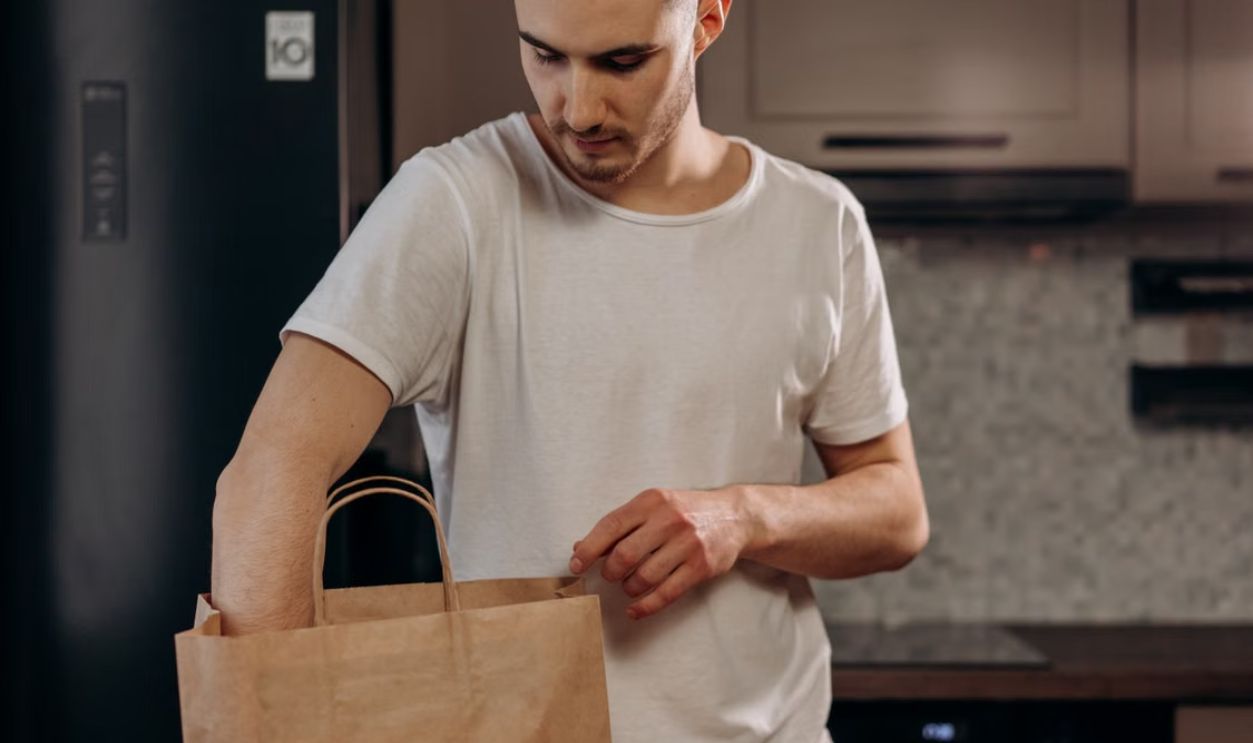 Male in white shirt unpacking groceries from paper bag in contemporary kitchen setting.