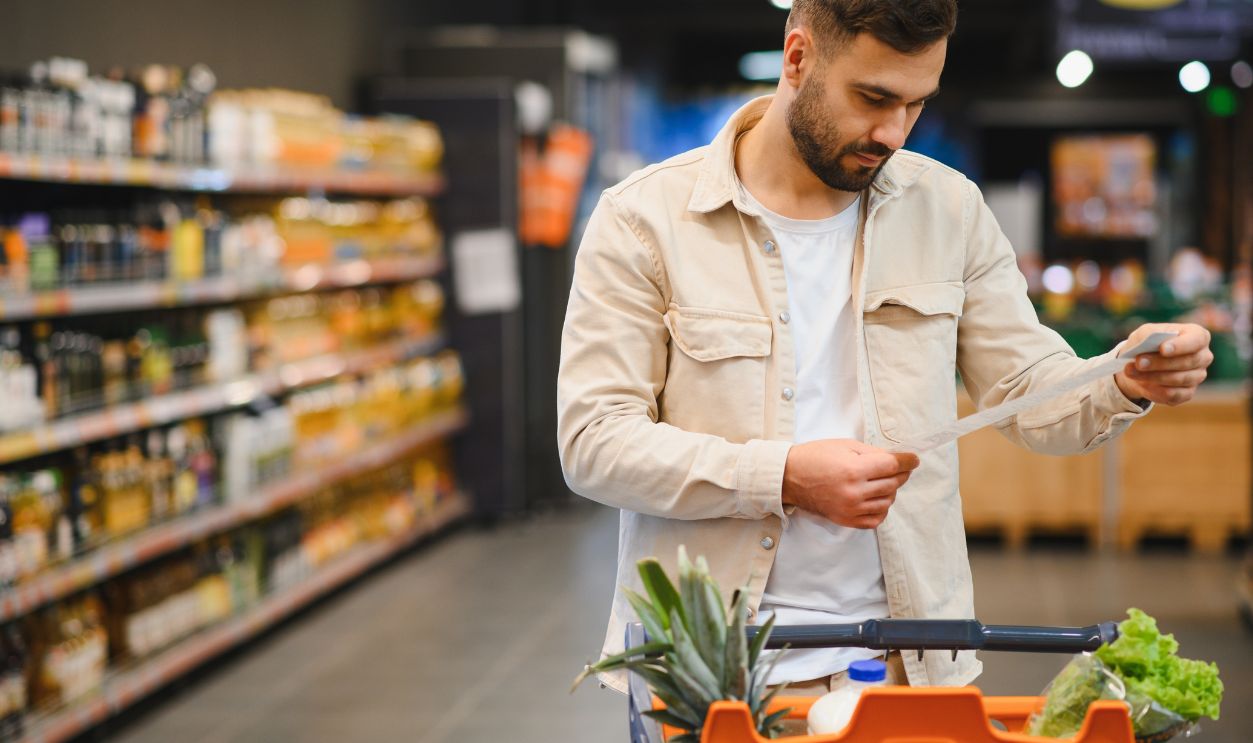 Shutterstock-2714234823, Man pushing grocery cart while carefully checking his shopping list in a supermarket aisle, focusing on budget and consumer choices