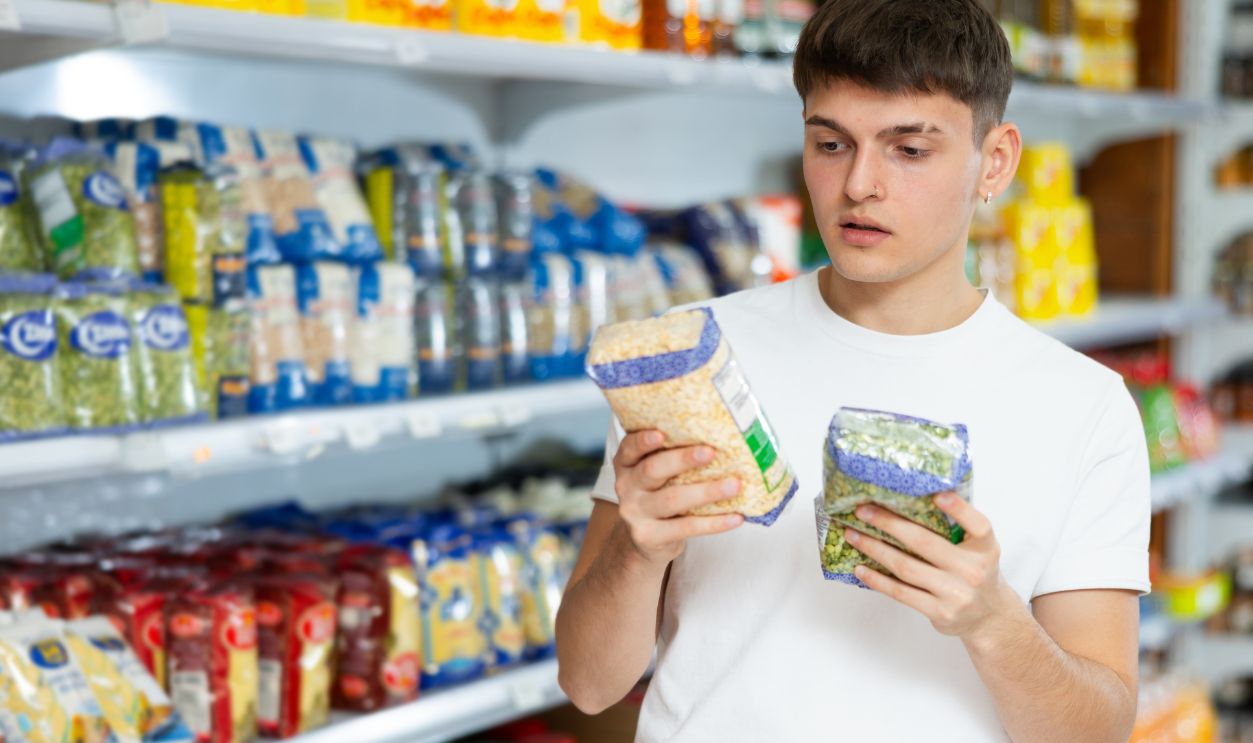 Portrait of pensive young guy choosing organic dried vegetables in supermarket, holding packages with green and yellow split peas in hands..