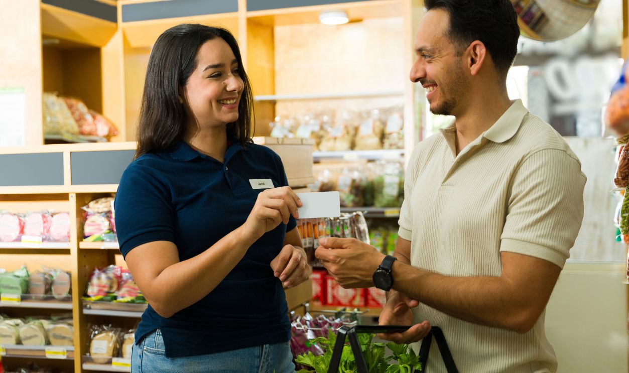 Shutterstock-2629167697, Cheerful supermarket employee giving loyalty card to male customer while smiling and talking in grocery store