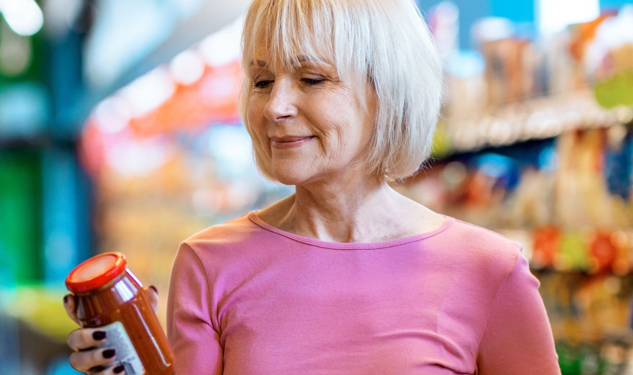 Senior Woman Shopping at Grocery Store While Holding a Jar of Sauce in Her Hand