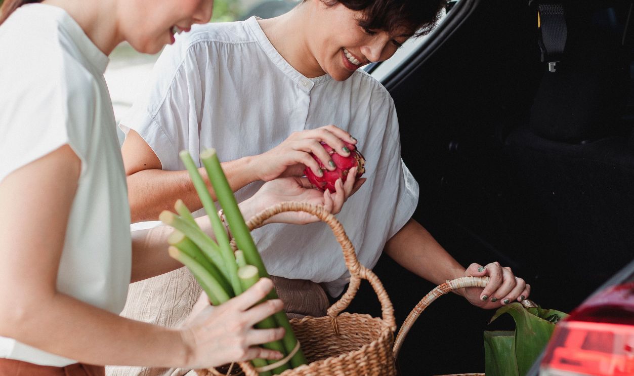 Two women loading fresh groceries into a car trunk, enjoying a sunny day outdoors