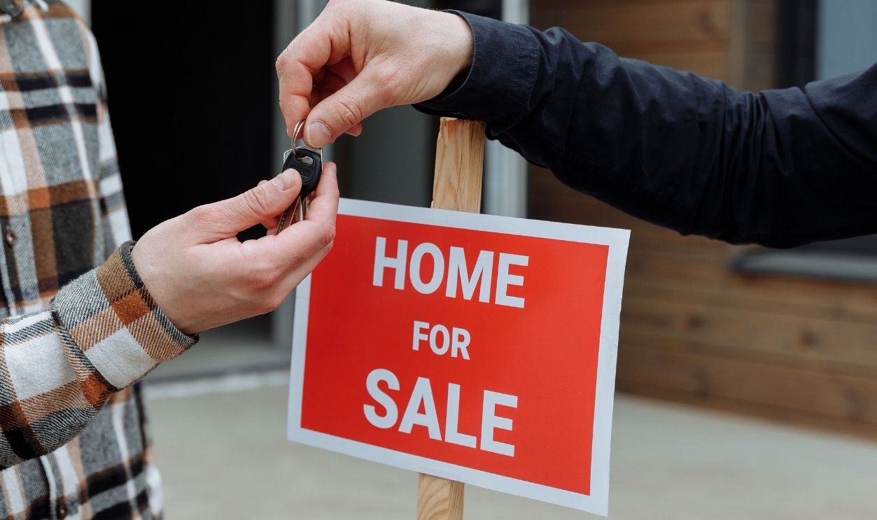 Close-up of a home for sale sign with people exchanging keys.