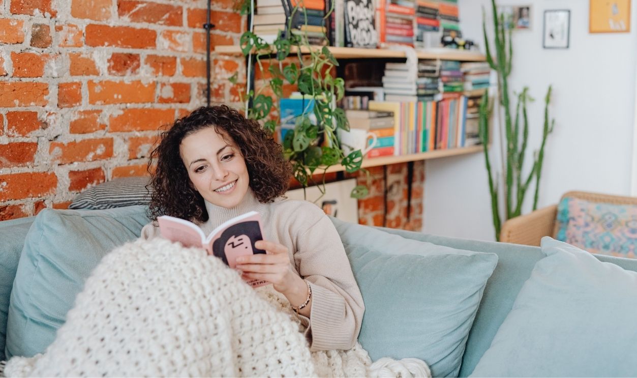 A smiling woman reads on a sofa in a cozy room with bookshelves and plants.