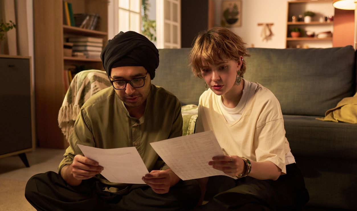 Two young adults sitting on floor, focused on reviewing bills and financial documents, demonstrating collaboration and discussion in a cozy living room environment