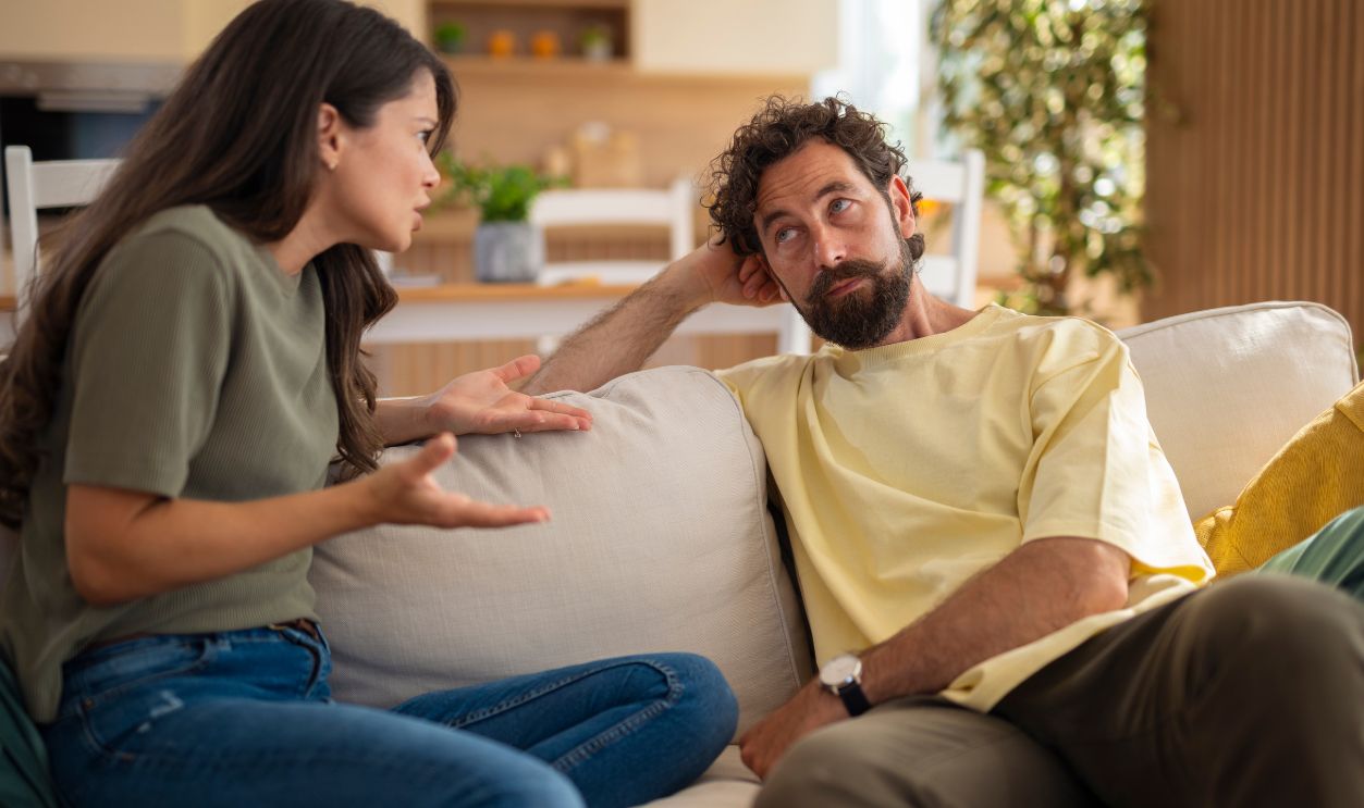 Couple having a serious conversation at home on the couch. Woman gesturing while man listens with a thoughtful expression. Bright natural light and modern home interior in the background.