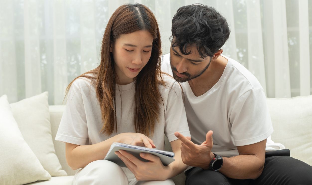 Young asian couple having serious discussion over finance document while sitting on comfortable sofa inside bright living room showing deep focus and concerned emotional state