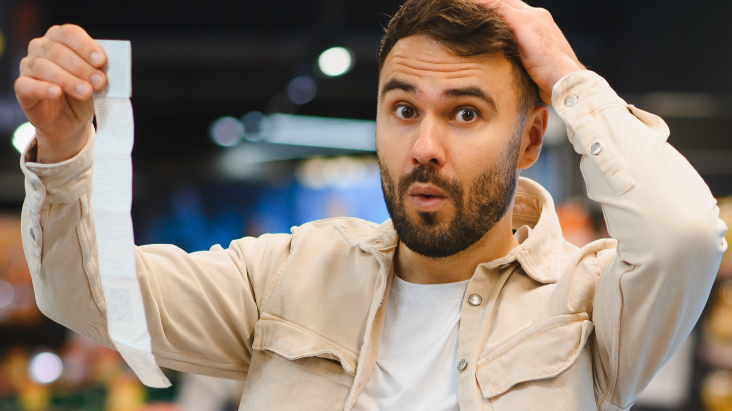 Man holding a long supermarket receipt, expressing shock and frustration about rising food costs