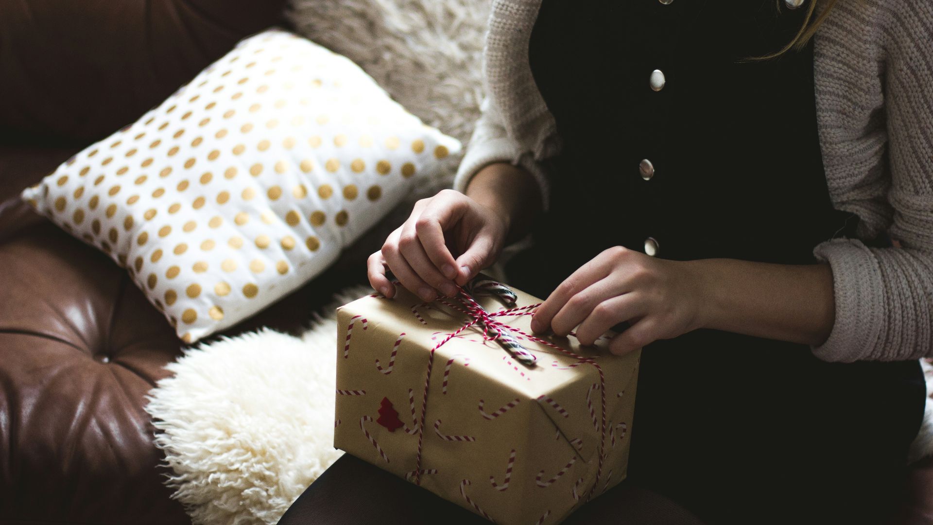 woman in grey sweater and blue denim dungarees sitting on brown and white sofa opening a yellow gift box closeup photography