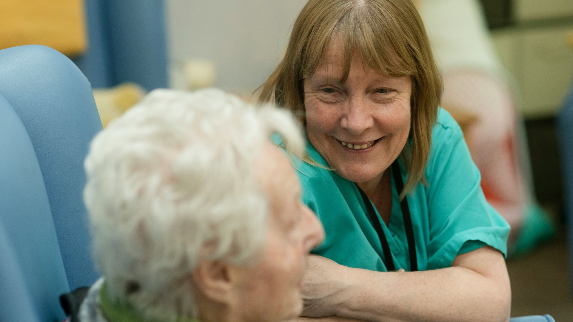 A smiling woman in green scrubs leaning toward an elderly person in a chair