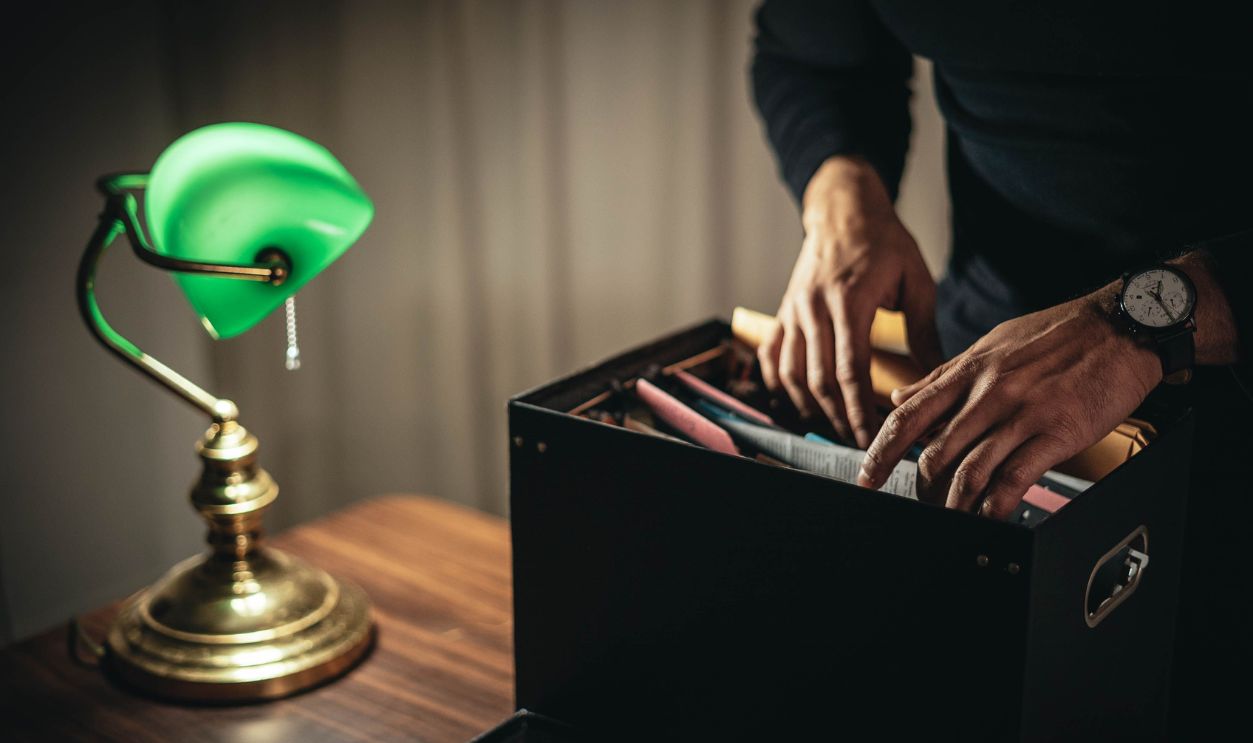 A person organizing documents in a file box under a green desk lamp on a wooden table