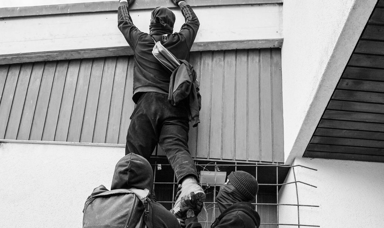 Three people in masks climbing a fence, suggesting trespassing in black and white.