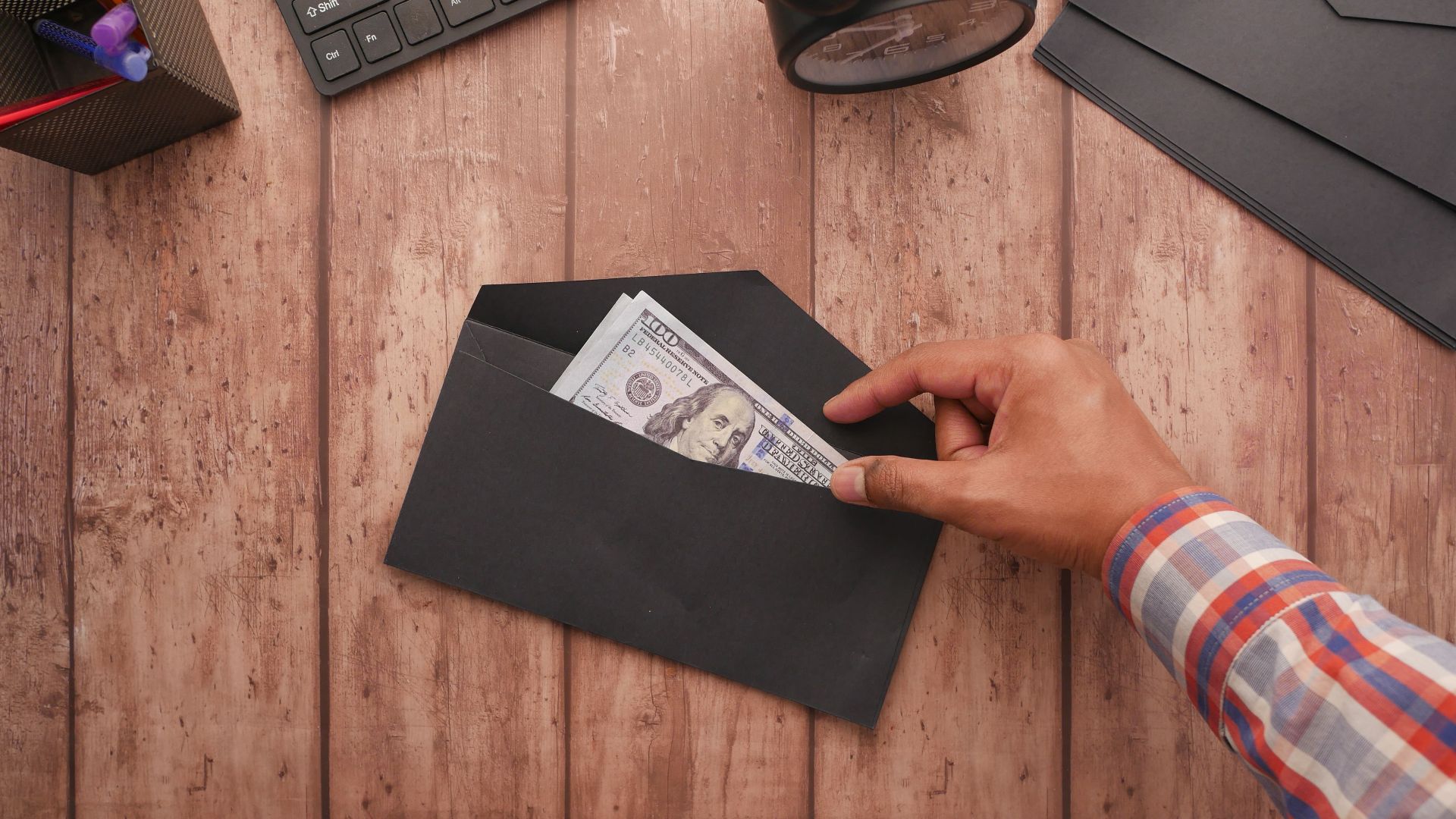A hand placing a 100 dollar bill in a black envelope on a wooden desk with stationery, keyboard, and alarm clock.