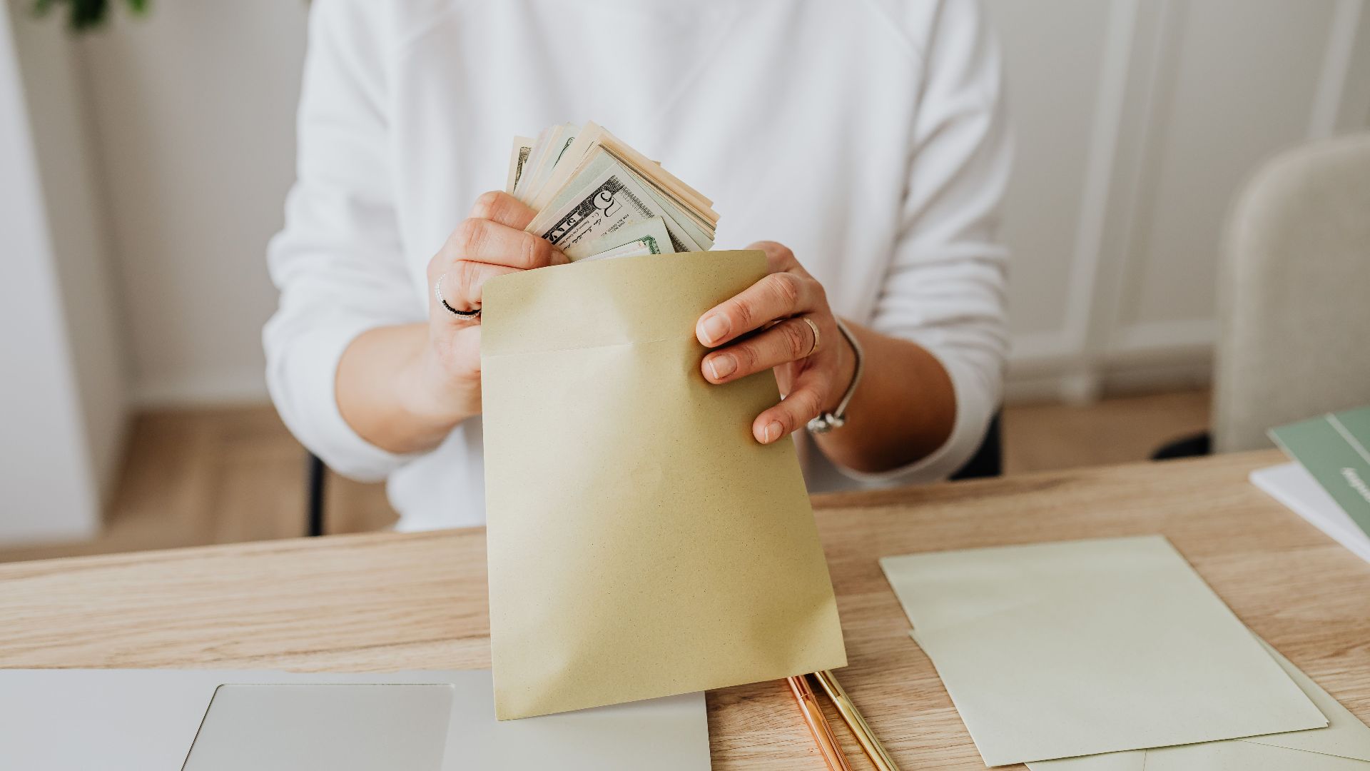 Close-up of person holding envelopes with cash at a wooden desk indoors.