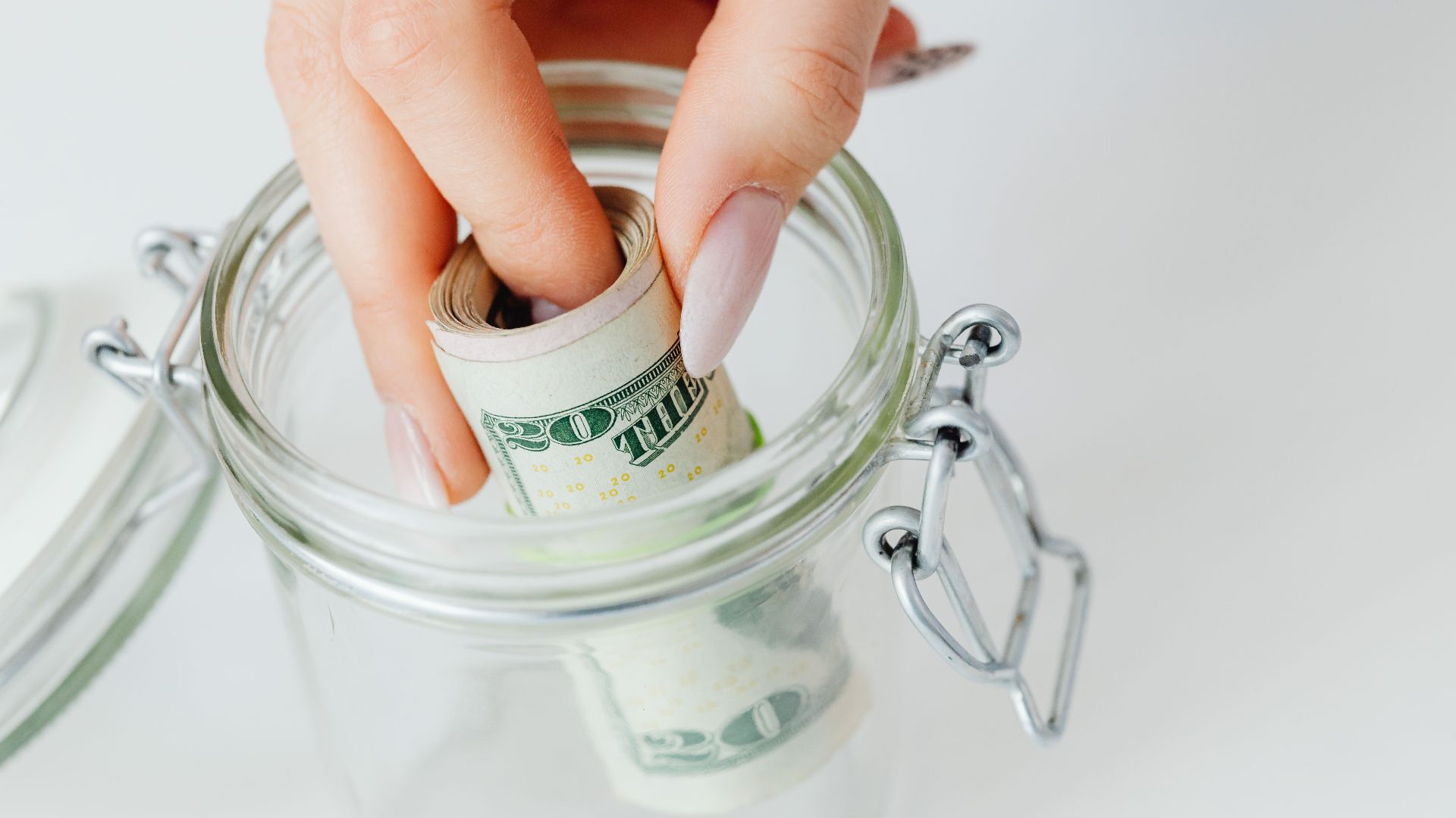 A close-up of a hand placing rolled dollars into a glass jar, symbolizing savings.