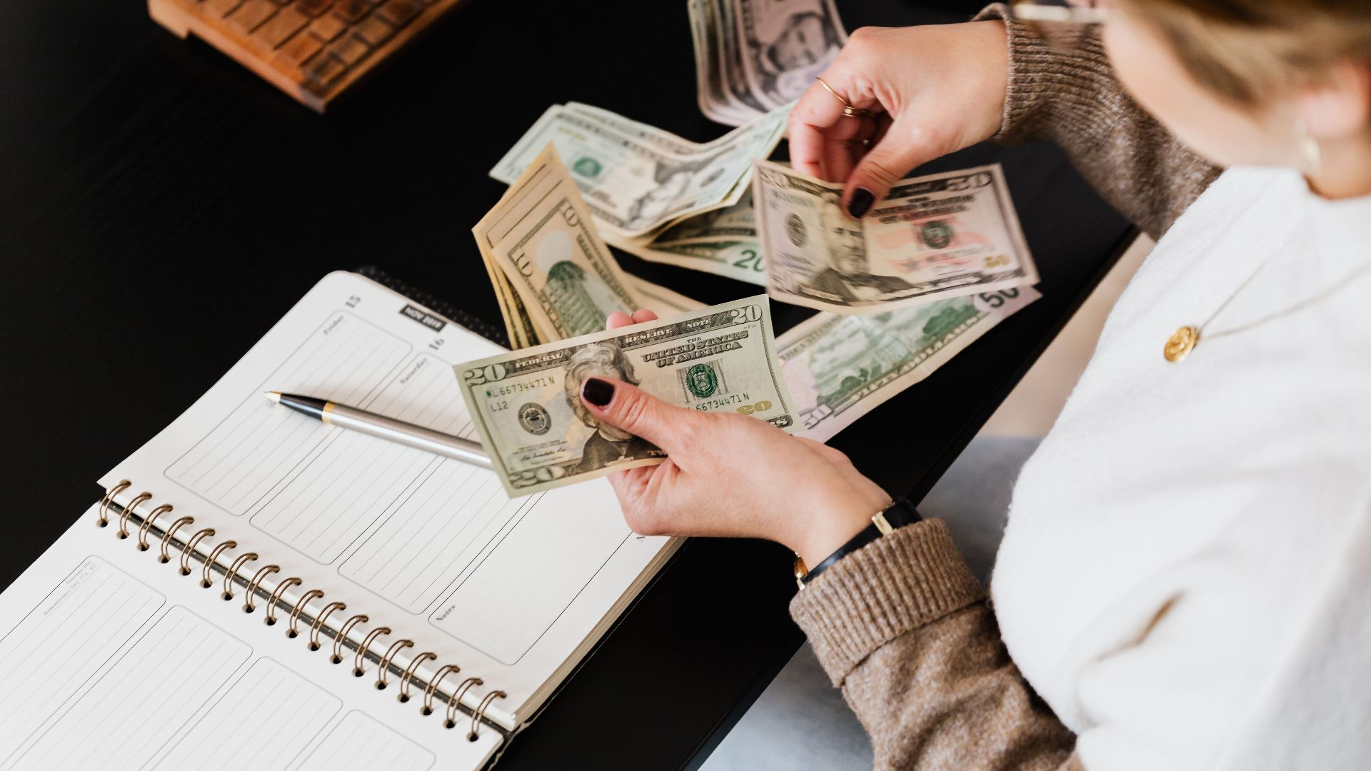 Woman organizing finances, counting dollar bills at desk with open notebook.