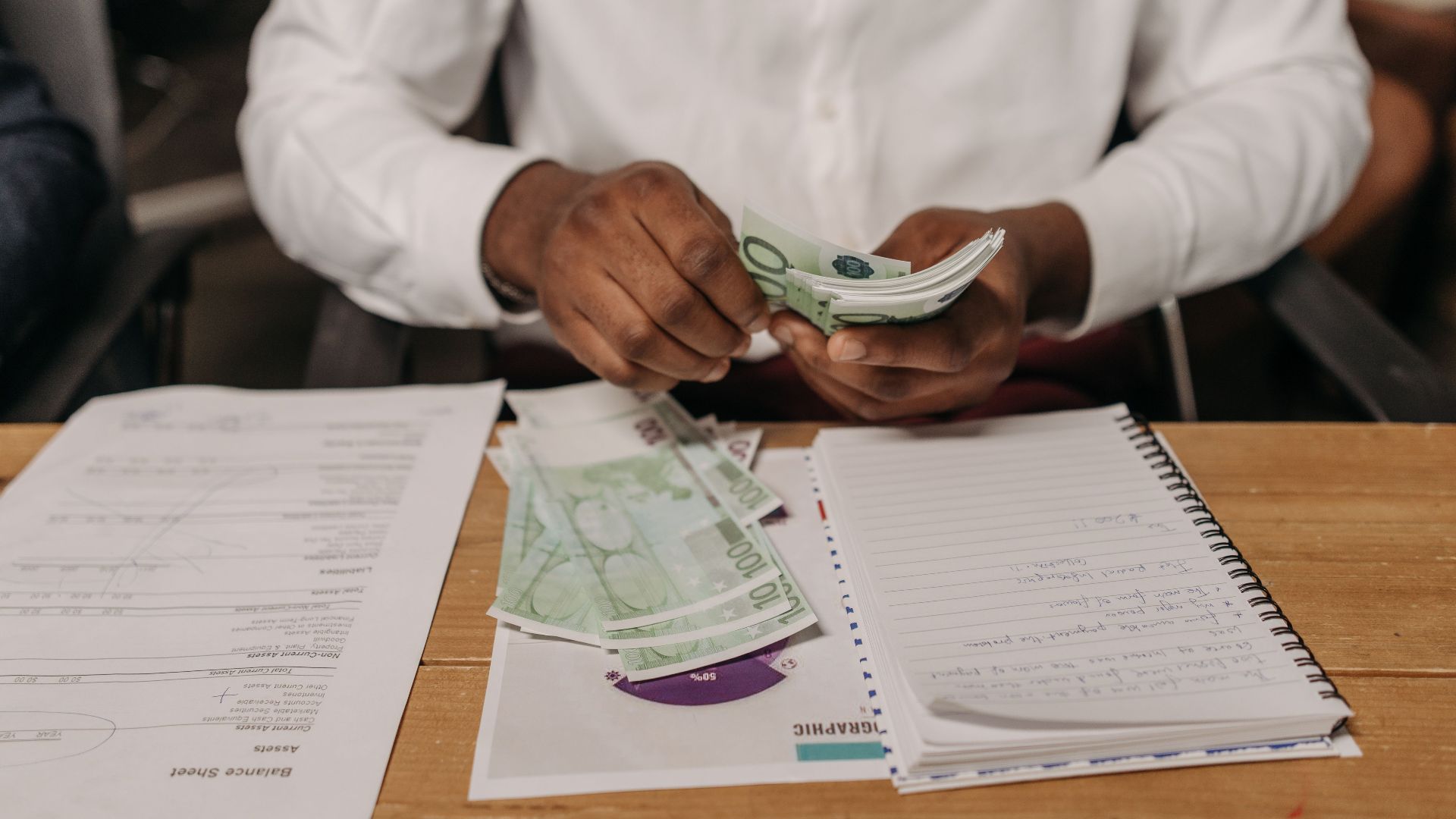 African businessman counting euro bills at a desk with documents and a notebook, highlighting finance theme.