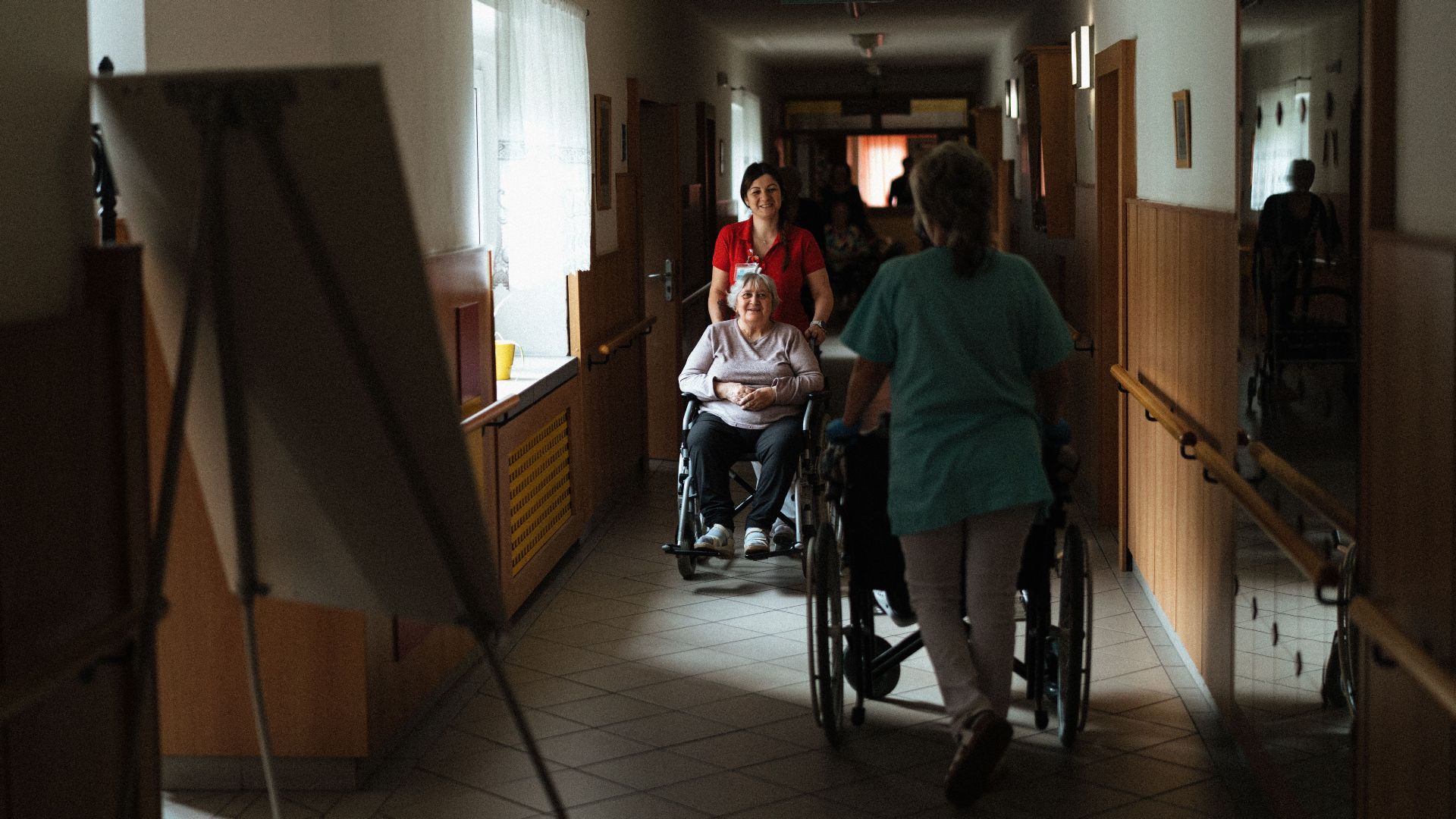 Caregiver assisting elderly woman in a wheelchair through a retirement home corridor.