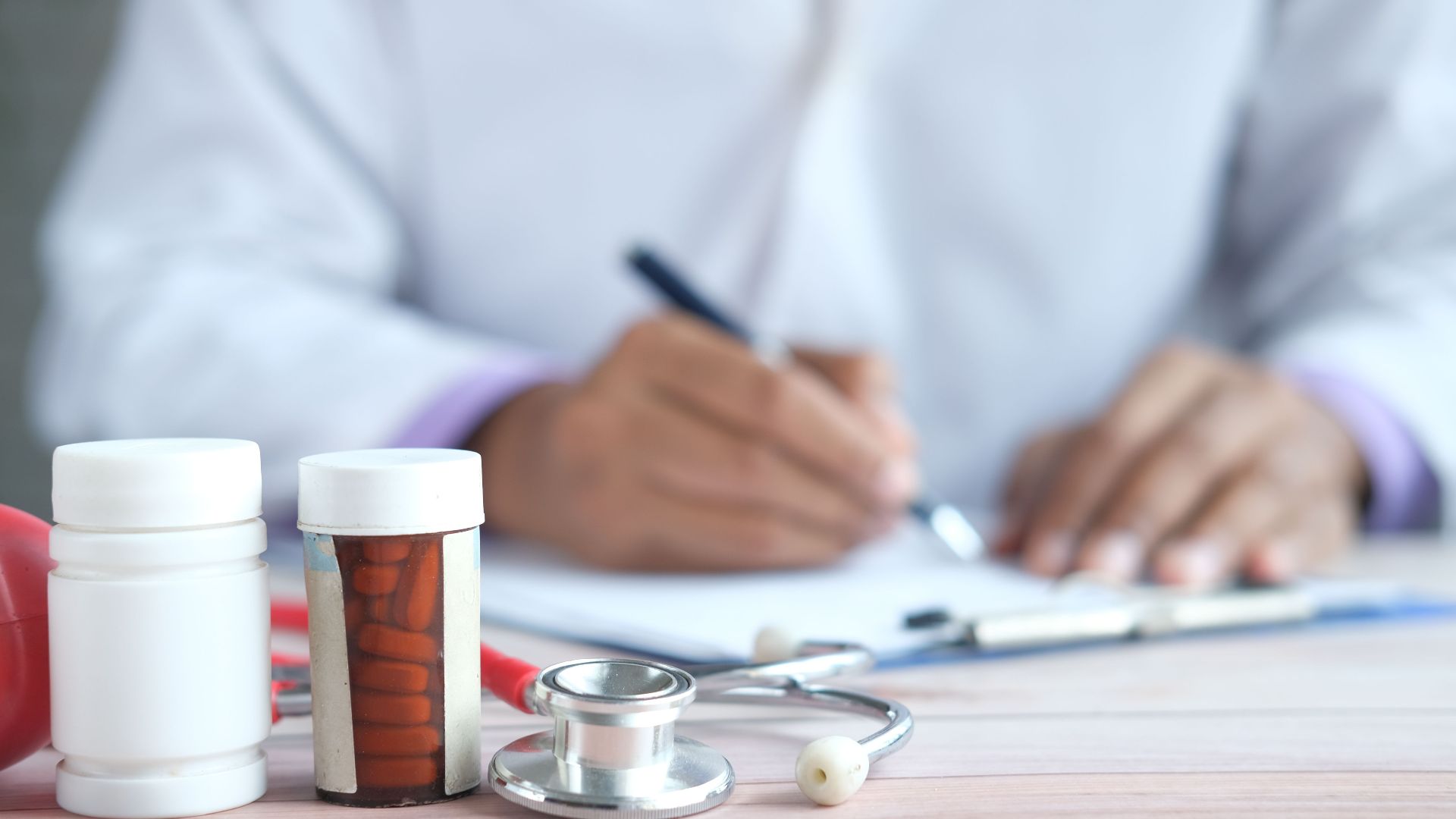 Close-up of doctor writing prescription with medicines and stethoscope on desk.