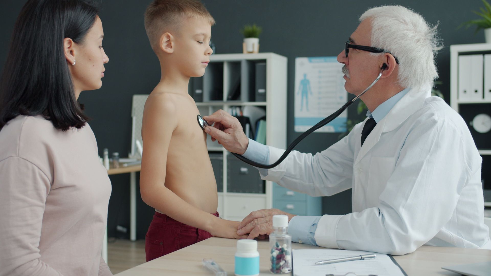 A doctor uses a stethoscope to examine a young boy with his mother present in a medical office.