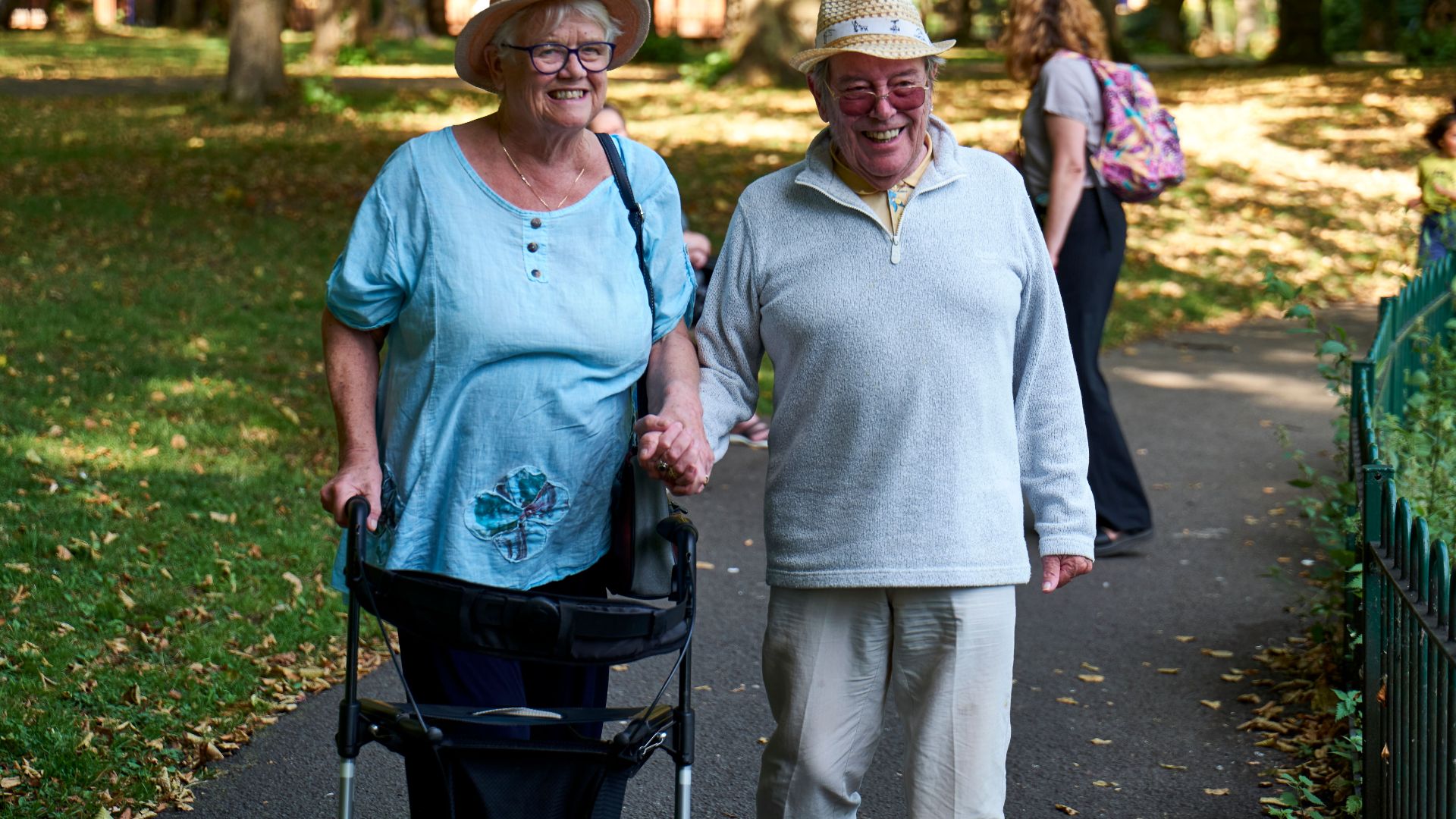 Cheerful elderly couple in casual clothes and straw hats walking on lawn of park in sunny day