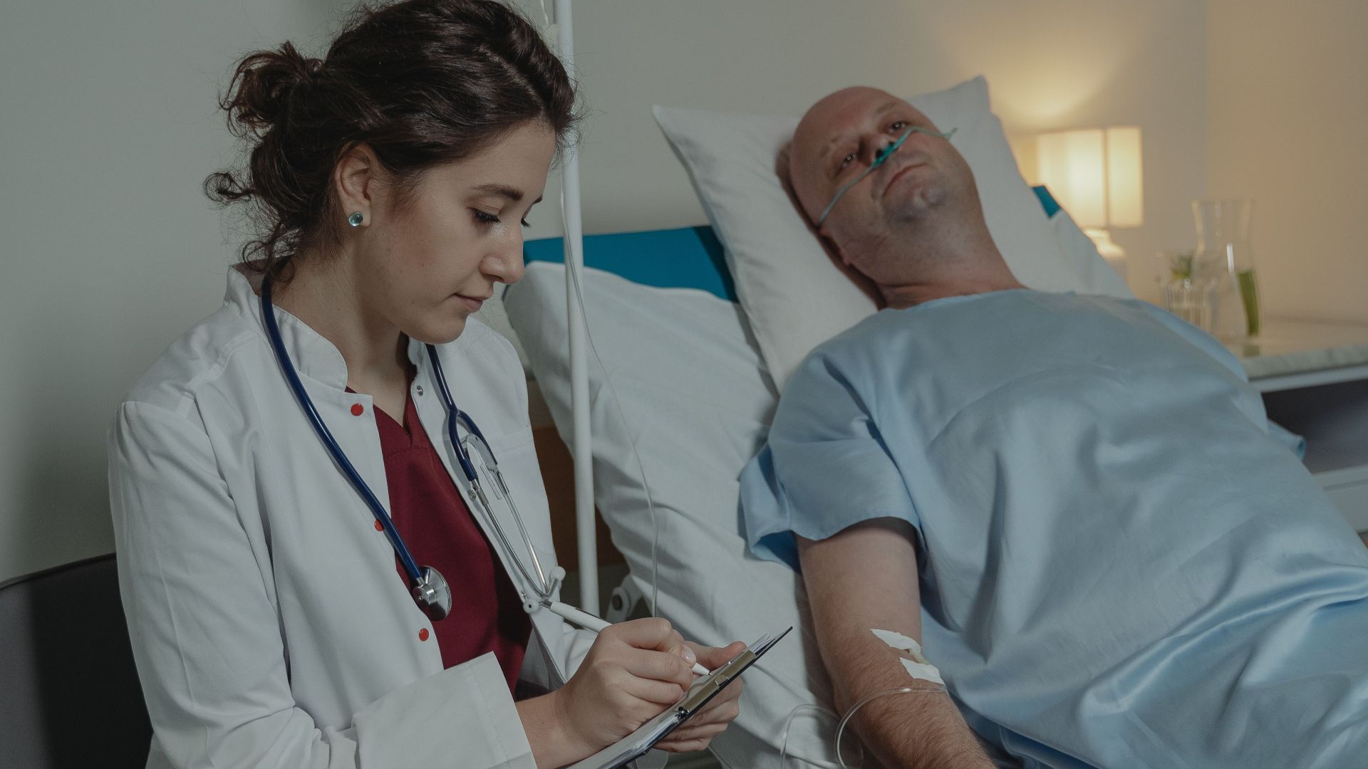 A doctor sits beside a patient in a hospital room, taking notes.