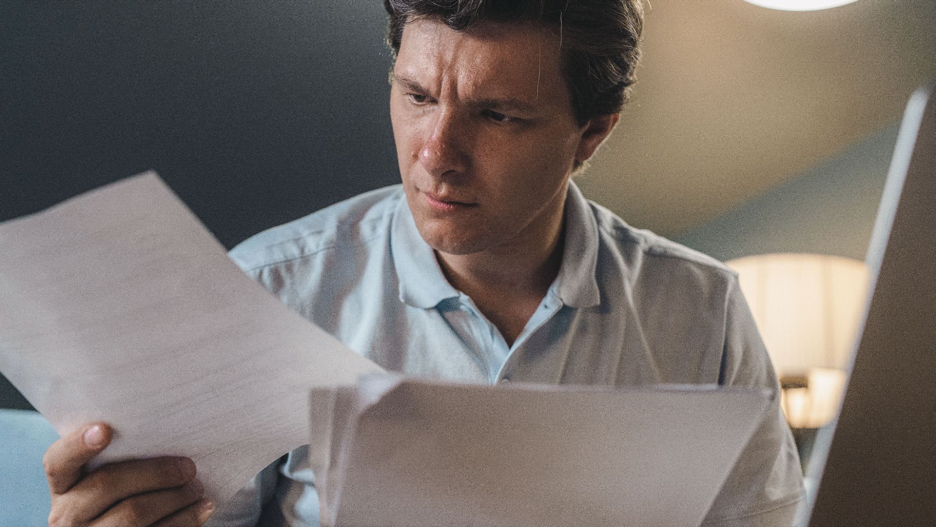 A man intently examines papers, seated indoors under warm lighting, focusing on his work.