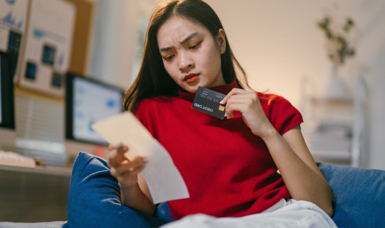 Young woman is sitting on a couch at home, looking worried and stressed as she checks her credit card bill and holds her credit card