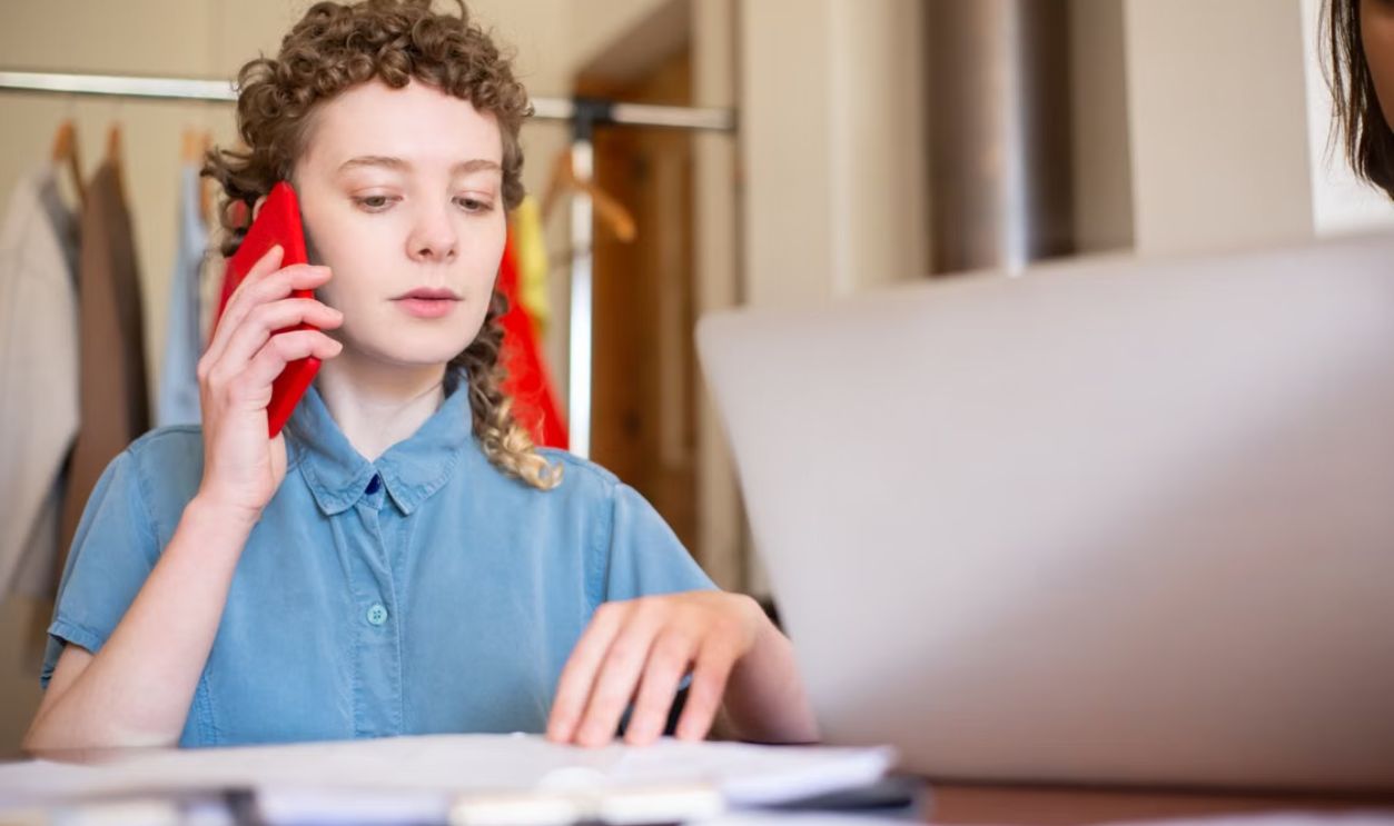 A woman with curly hair talks on the phone while working at a desk, highlighting a remote work lifestyle