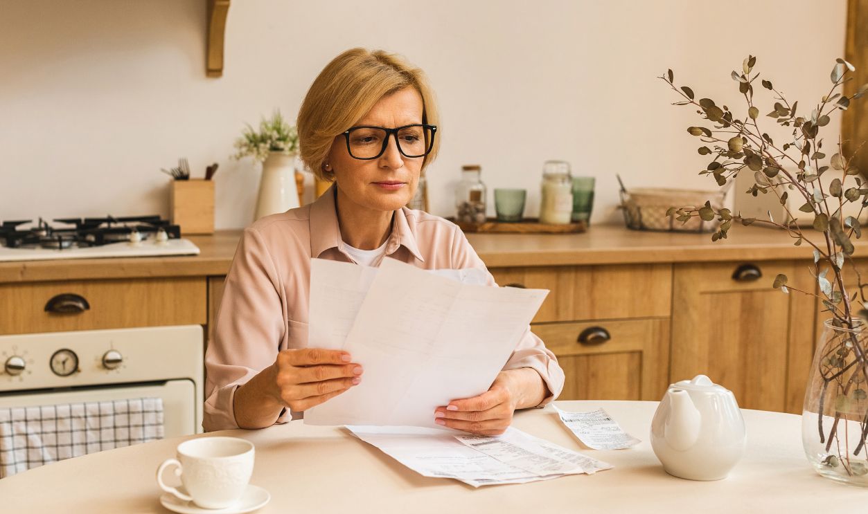 Middle aged mature senior woman holding paper bill or letter at home for making online payments on website, calculating financial taxes fee cost, reviewing bank account.