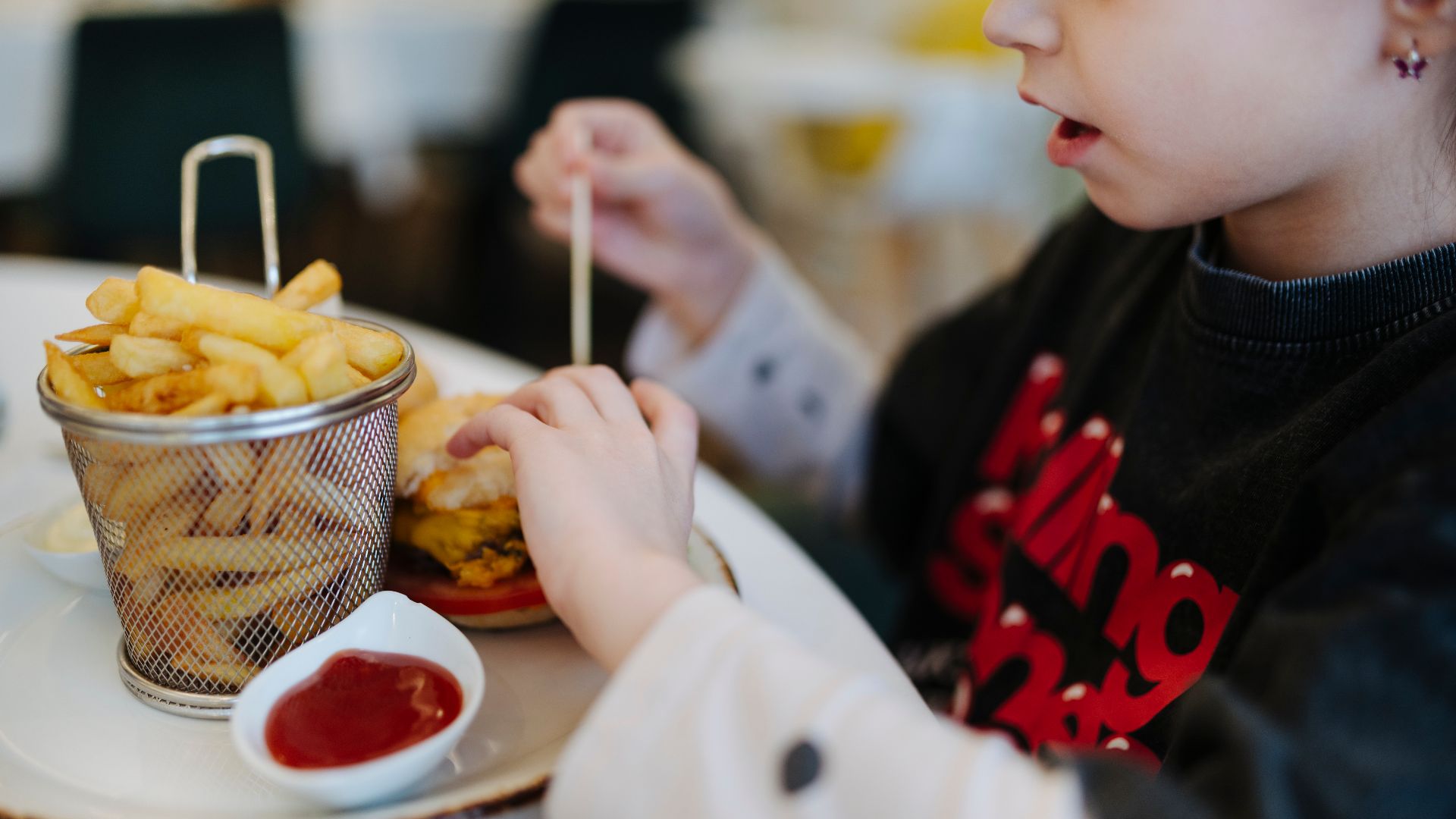A child sits at a table in a restaurant holding a burger. Next to the burger is a small basket of fries and a dish of ketchup. The scene shows food setup during lunchtime.