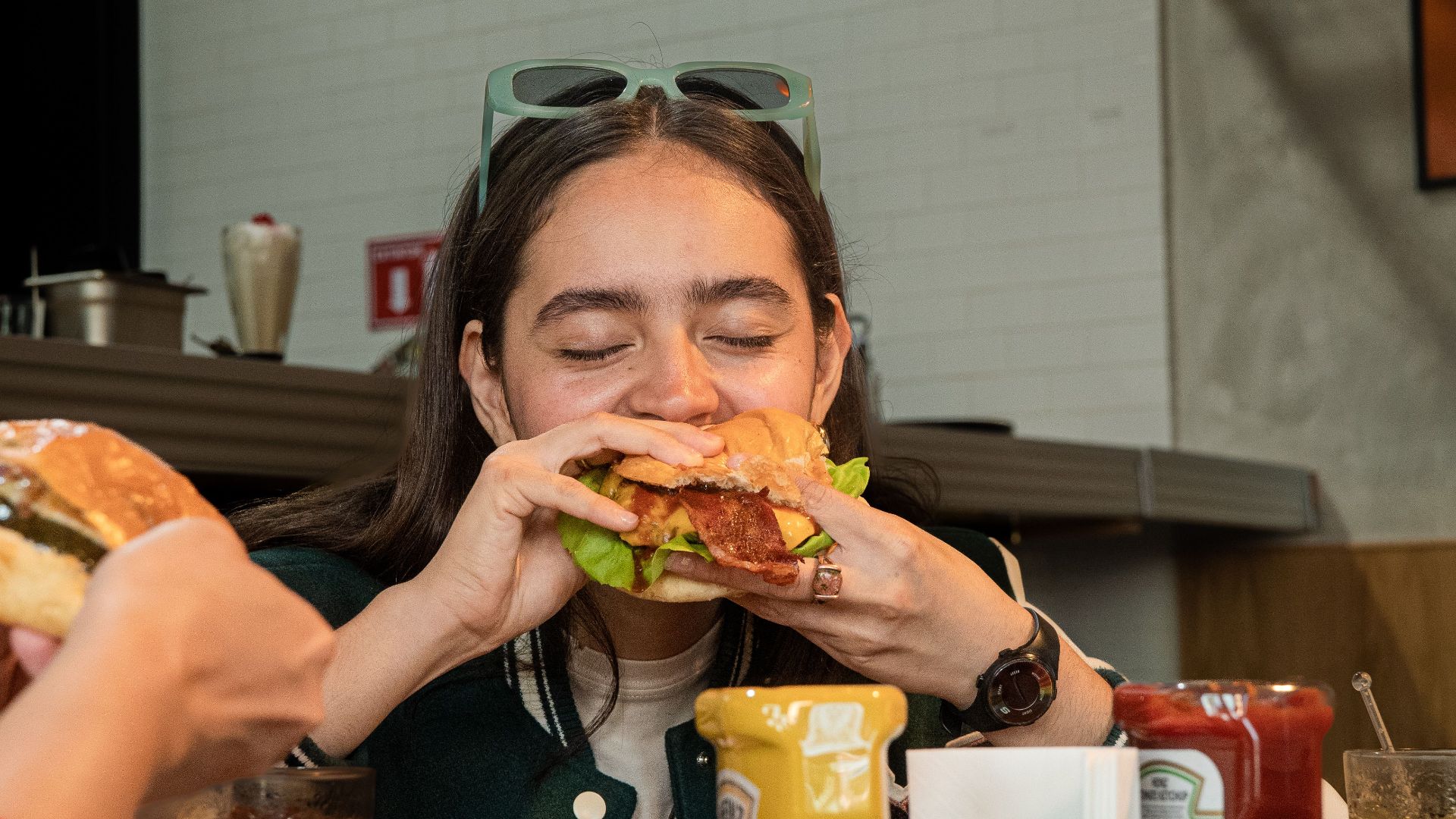 A young woman devours a juicy burger in a casual restaurant setting, surrounded by condiments.