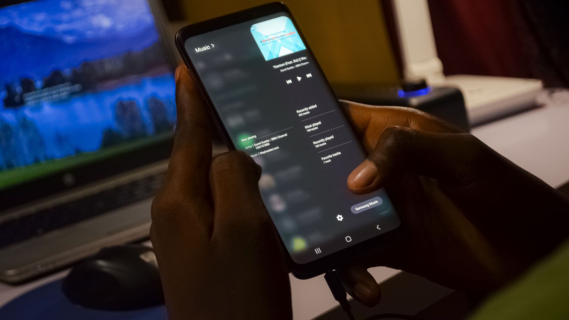 Person using smartphone with music app, indoors with laptop in background.